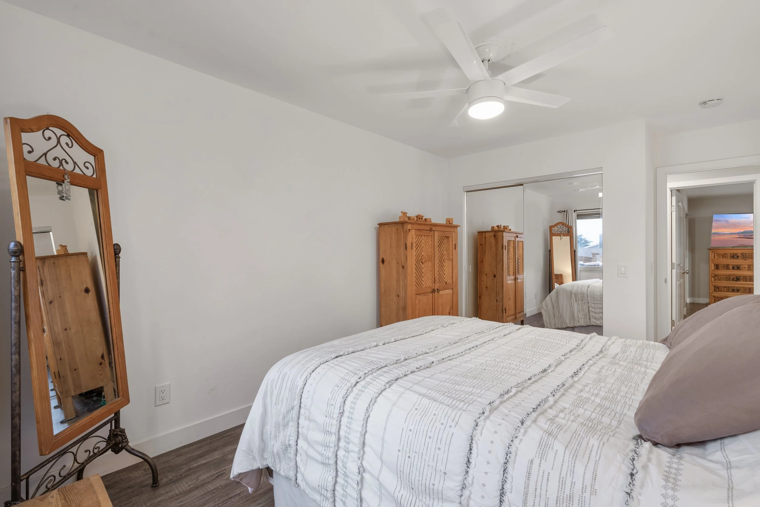 A bedroom with white walls, wooden furniture including a wardrobe and a tall mirror, a bed with white and beige bedding, a ceiling fan with lights, and a window showing an outside view.