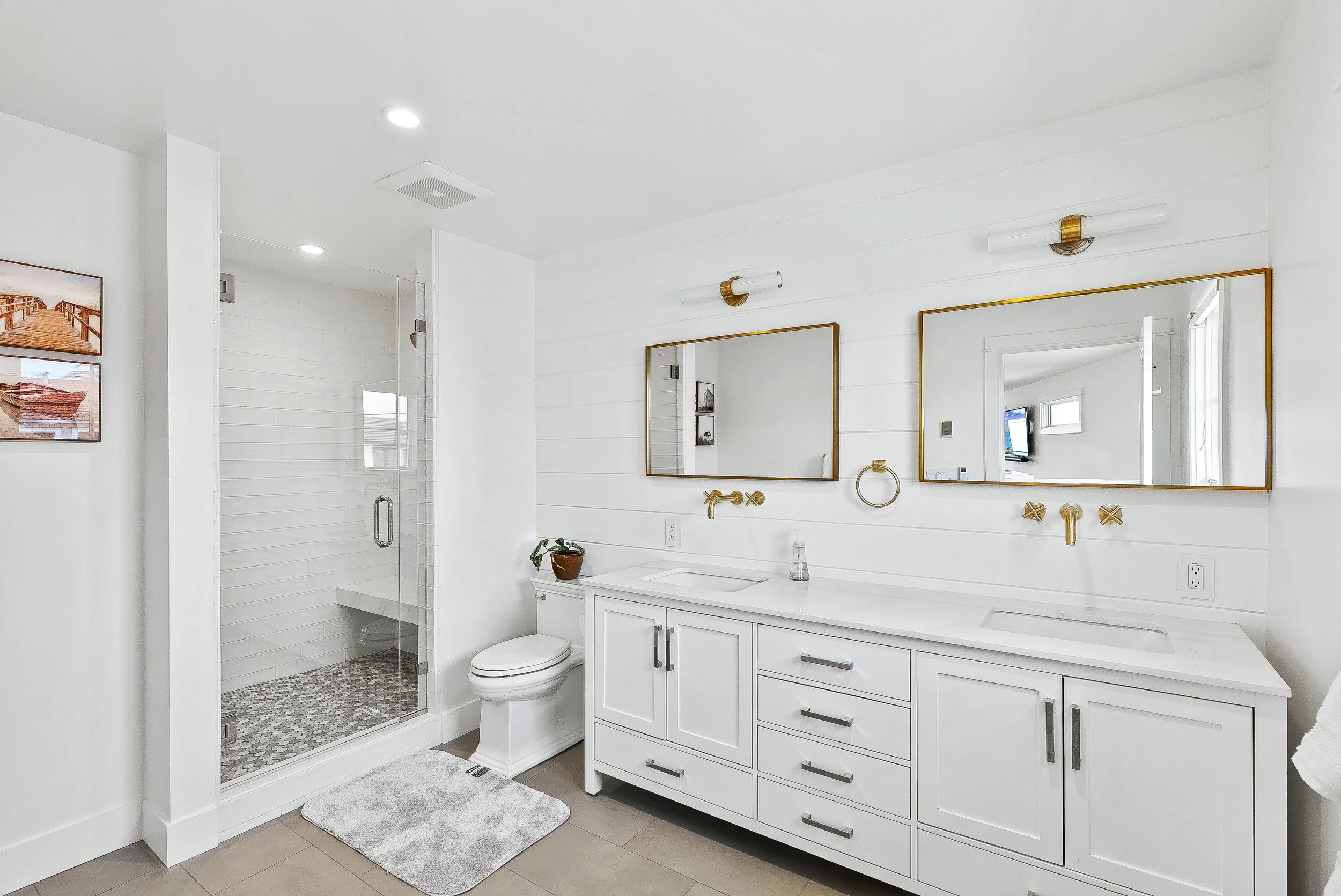 Modern white bathroom with a double vanity, two mirrors with gold frames, a walk-in shower with glass door, and a toilet. The room has wall-mounted lights, a potted plant, and a gray rug in Capitola, California.