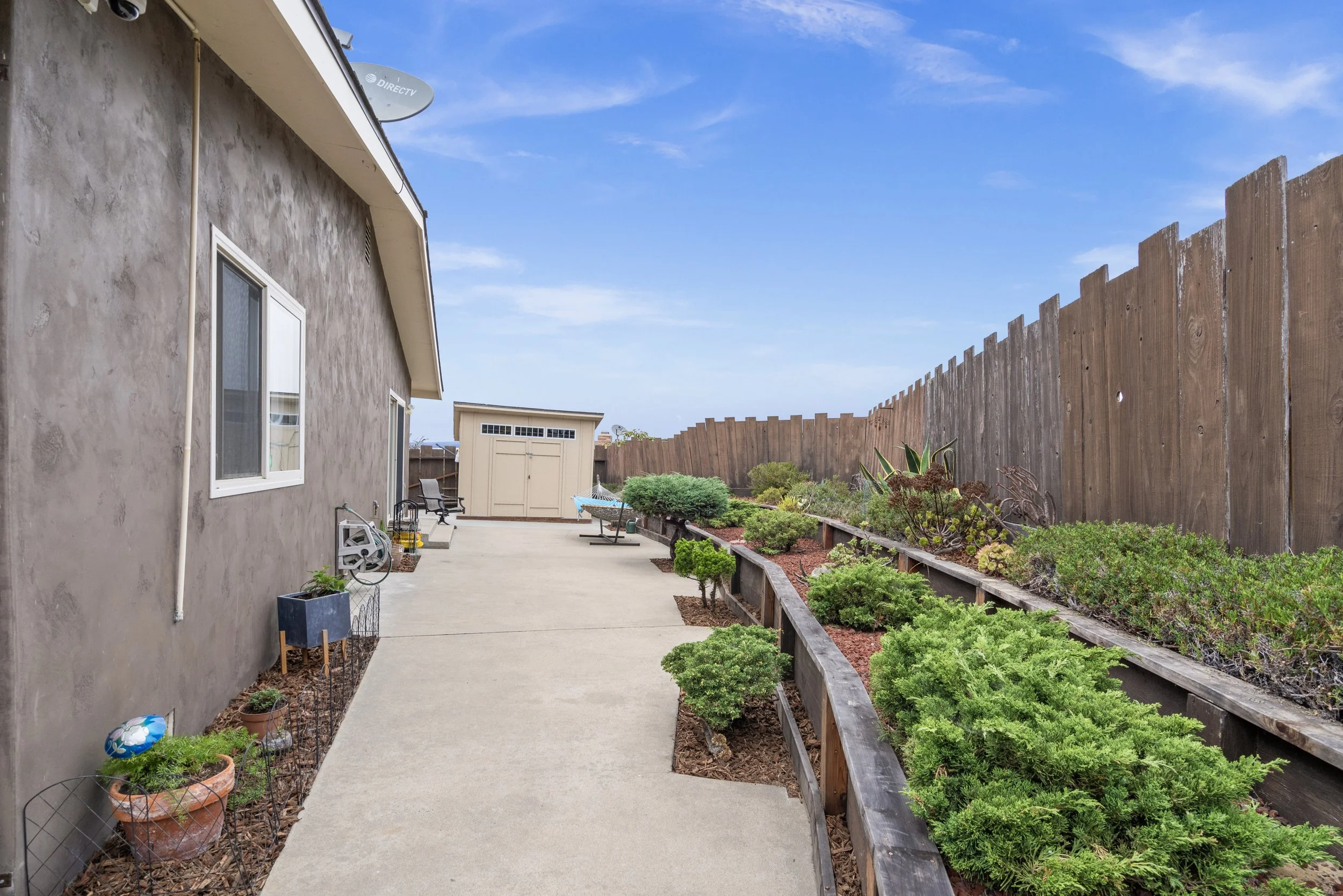 Backyard patio with concrete pathway, garden bed with various plants, shed, and wooden fence under a blue sky.