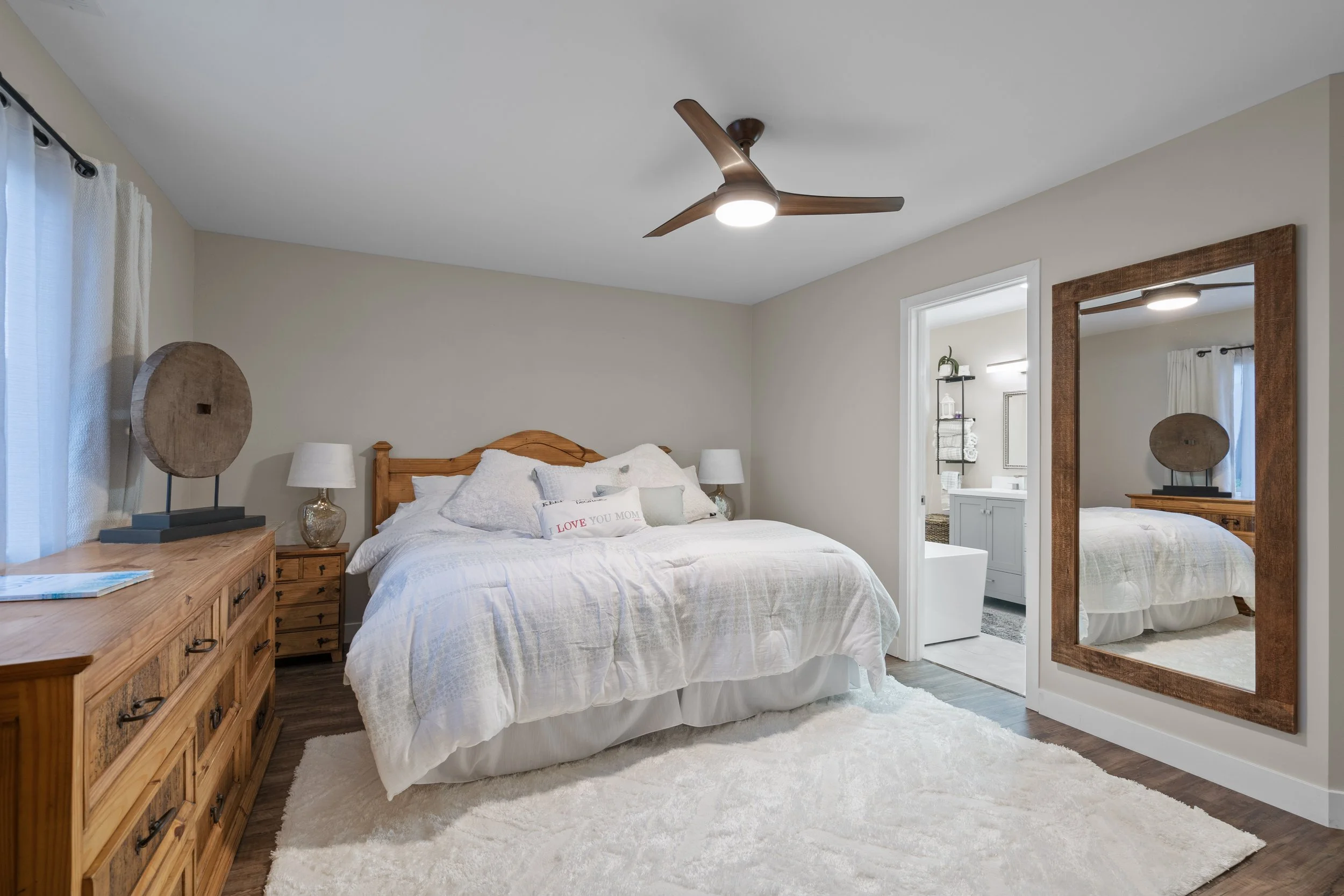 A bedroom with a wooden bed frame, white bedding, and multiple pillows, one with a message, flanked by nightstands with lamps, a wooden dresser, a large wall mirror, a ceiling fan, and a view into a bathroom with a vanity.