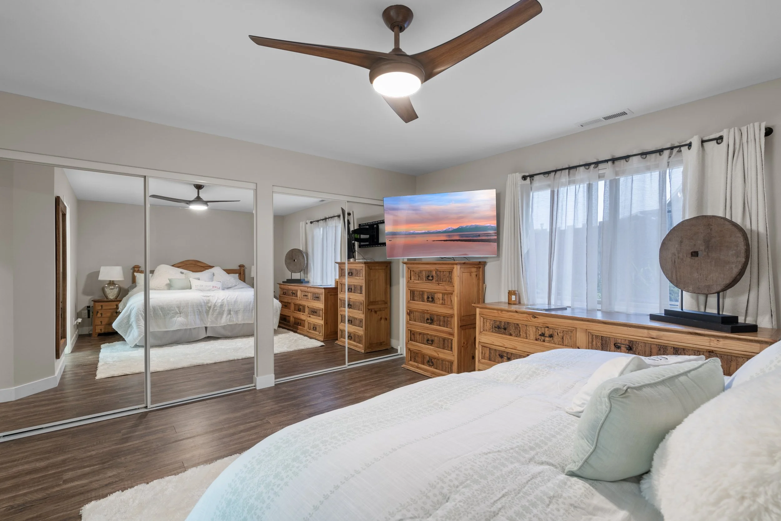 Bedroom with large bed, wooden furniture, mirrored closet doors, wall-mounted TV, ceiling fan, and window with light curtains