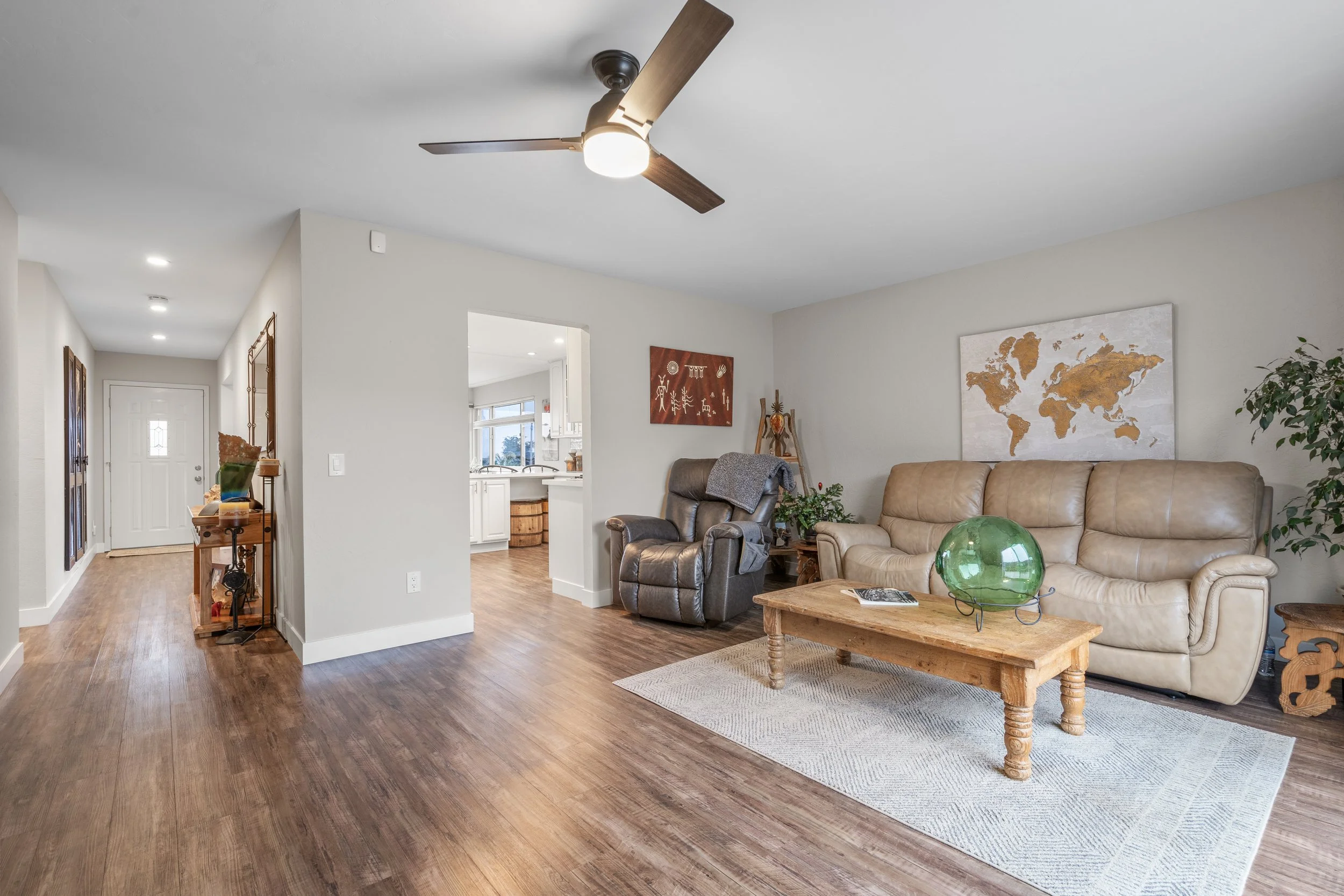 Living room with beige and brown furniture, world map wall art, ceiling fan, wooden coffee table with green glass decor, hardwood floors, and open view into a kitchen.