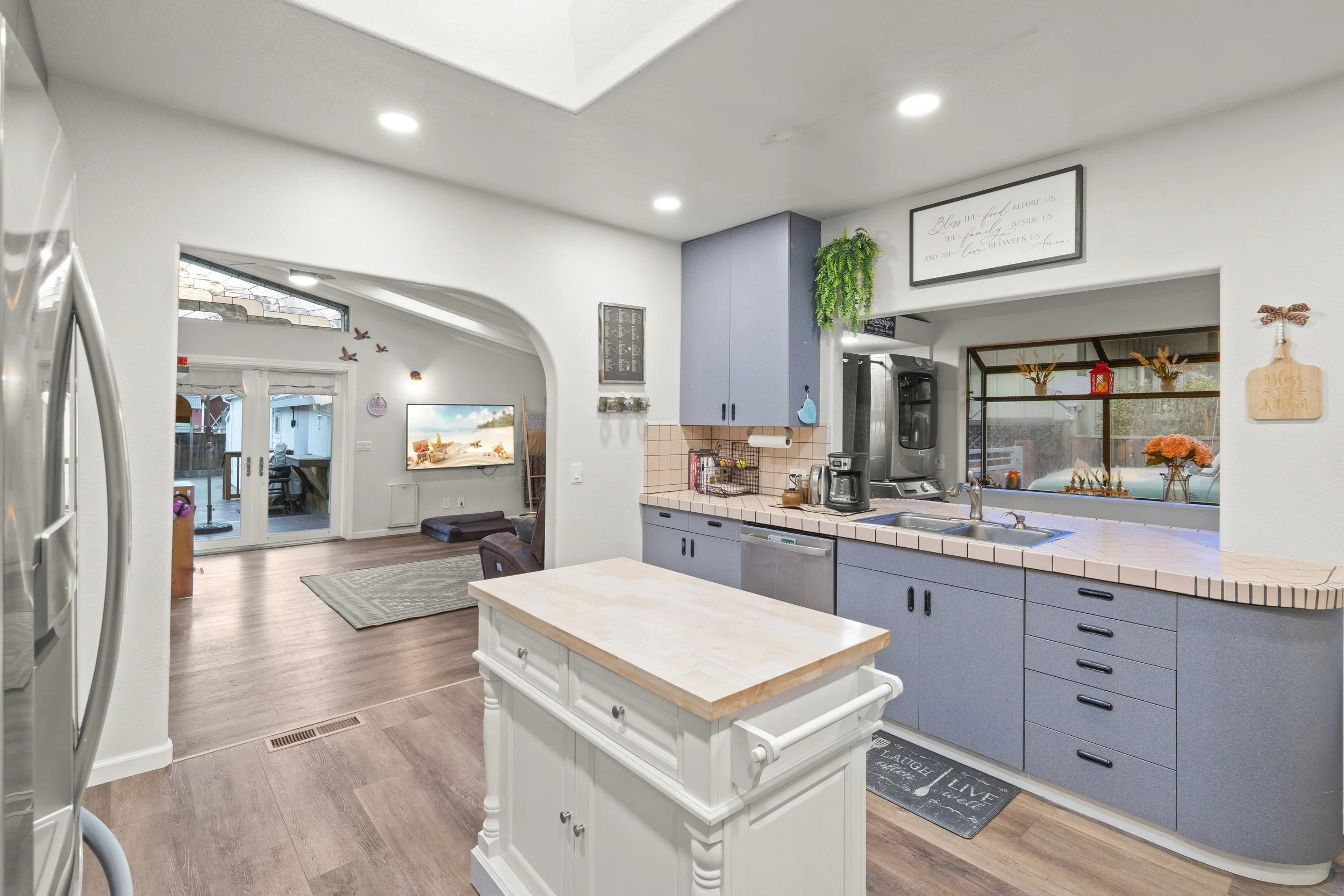Kitchen with blue cabinets, pink tiled countertops, and a window above the sink. Visible living room with TV and patio doors in the background.