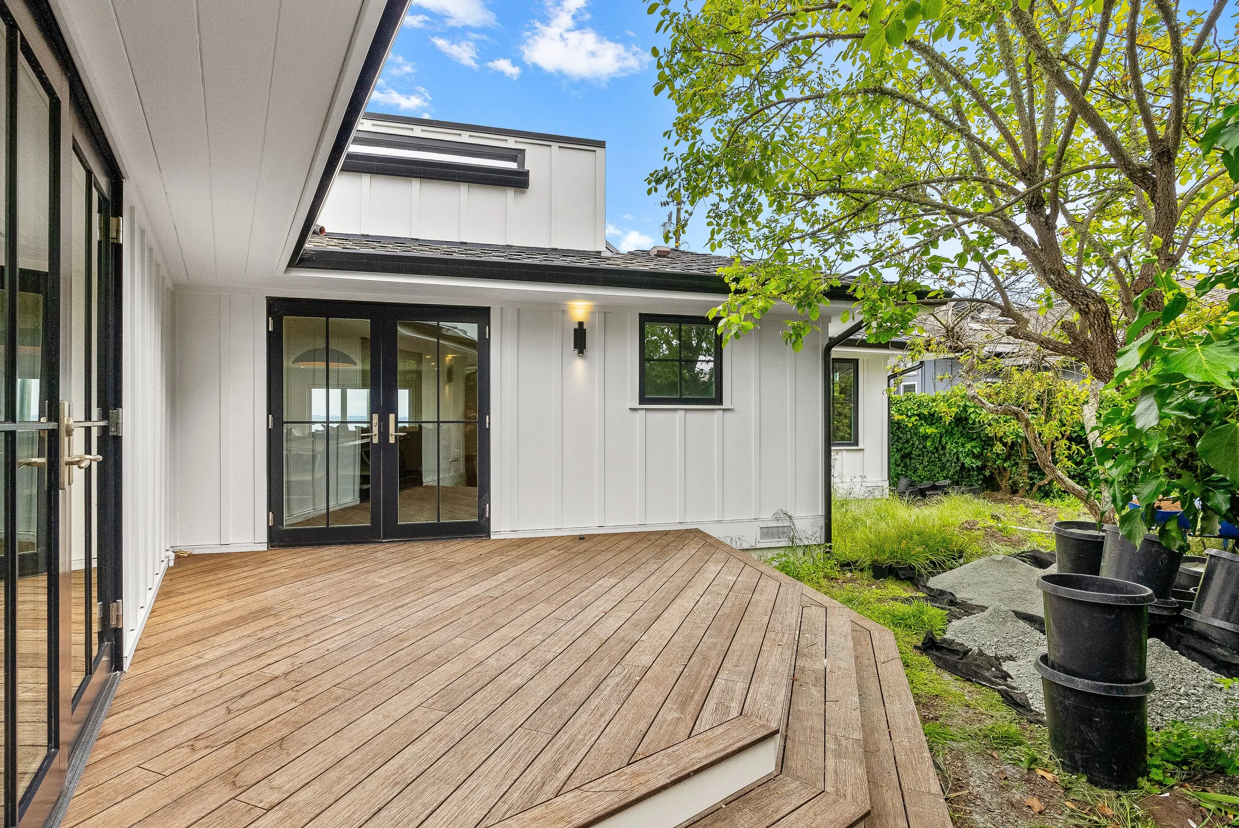 Backyard patio with a wooden deck, white exterior house, black framed glass doors, and a tree with green leavesin Capitola, California.