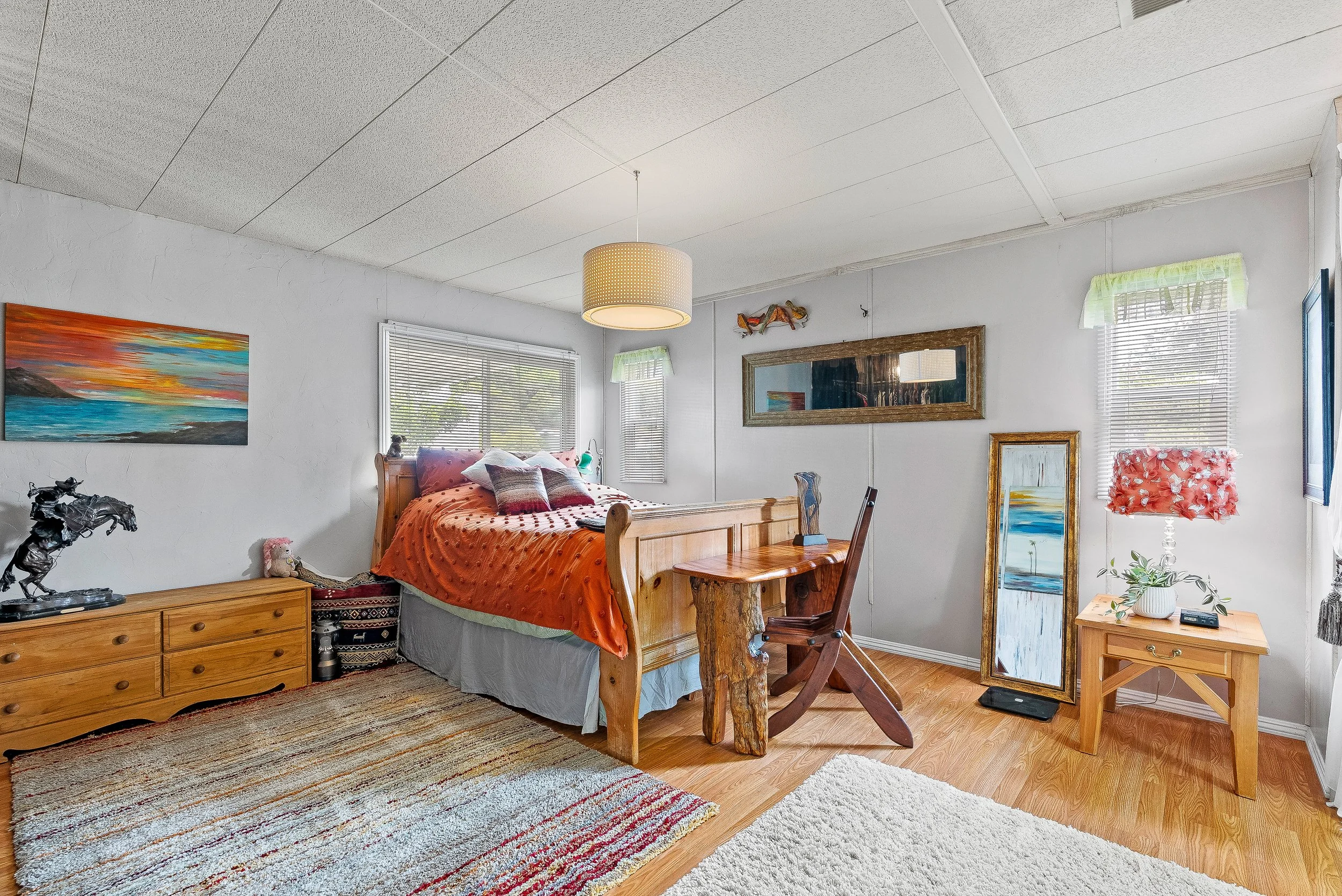 A bedroom with a wooden bed, orange bedspread, colorful pillows, and various artworks on white walls, with windows letting in natural light, a wooden dresser, a small wooden table, a mirror, and a colorful table lamp in Capitola, California.