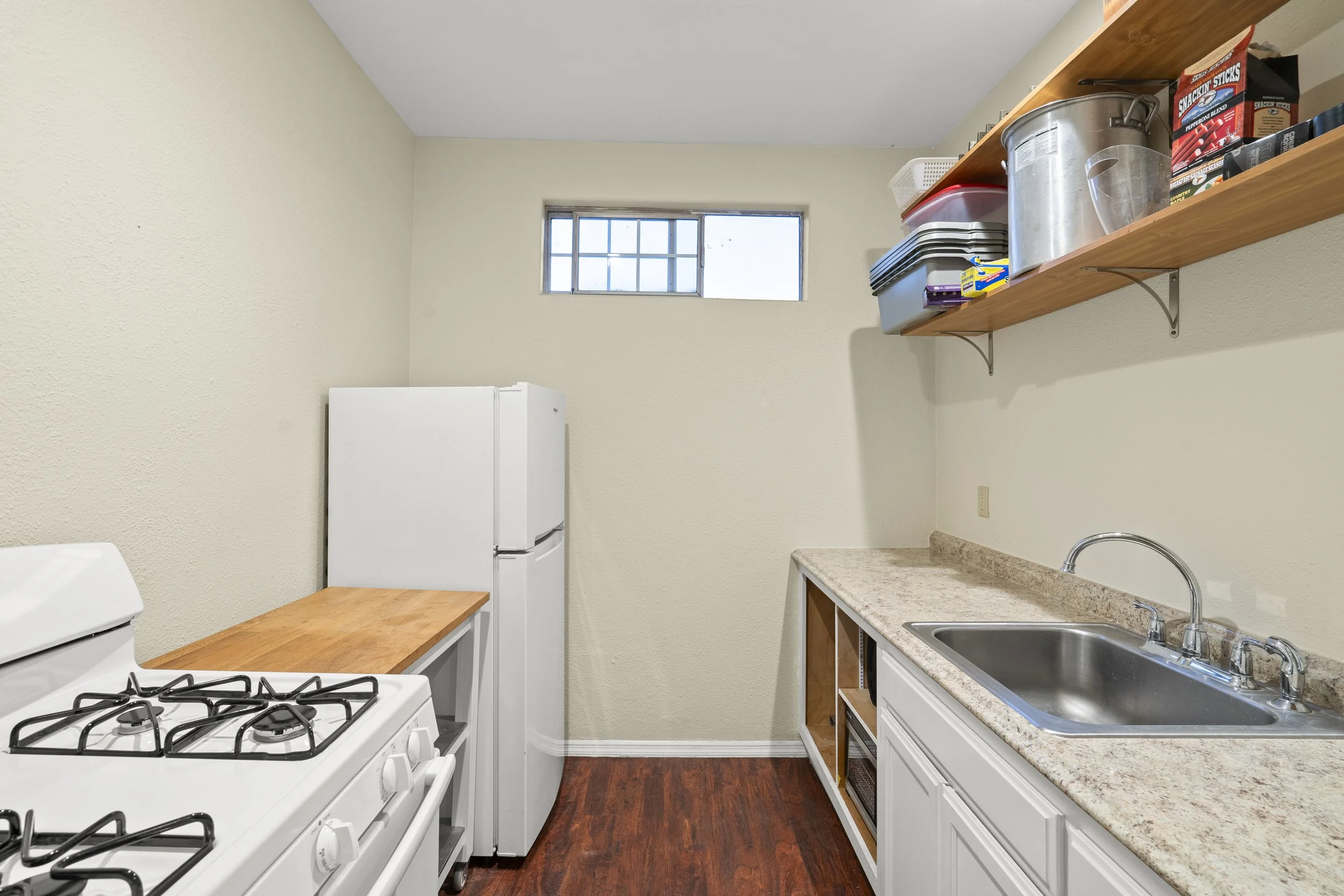 Small kitchen with white stove, refrigerator, beige countertop, stainless steel sink, open shelving with containers and food items, window with bars, wood floor, beige walls.