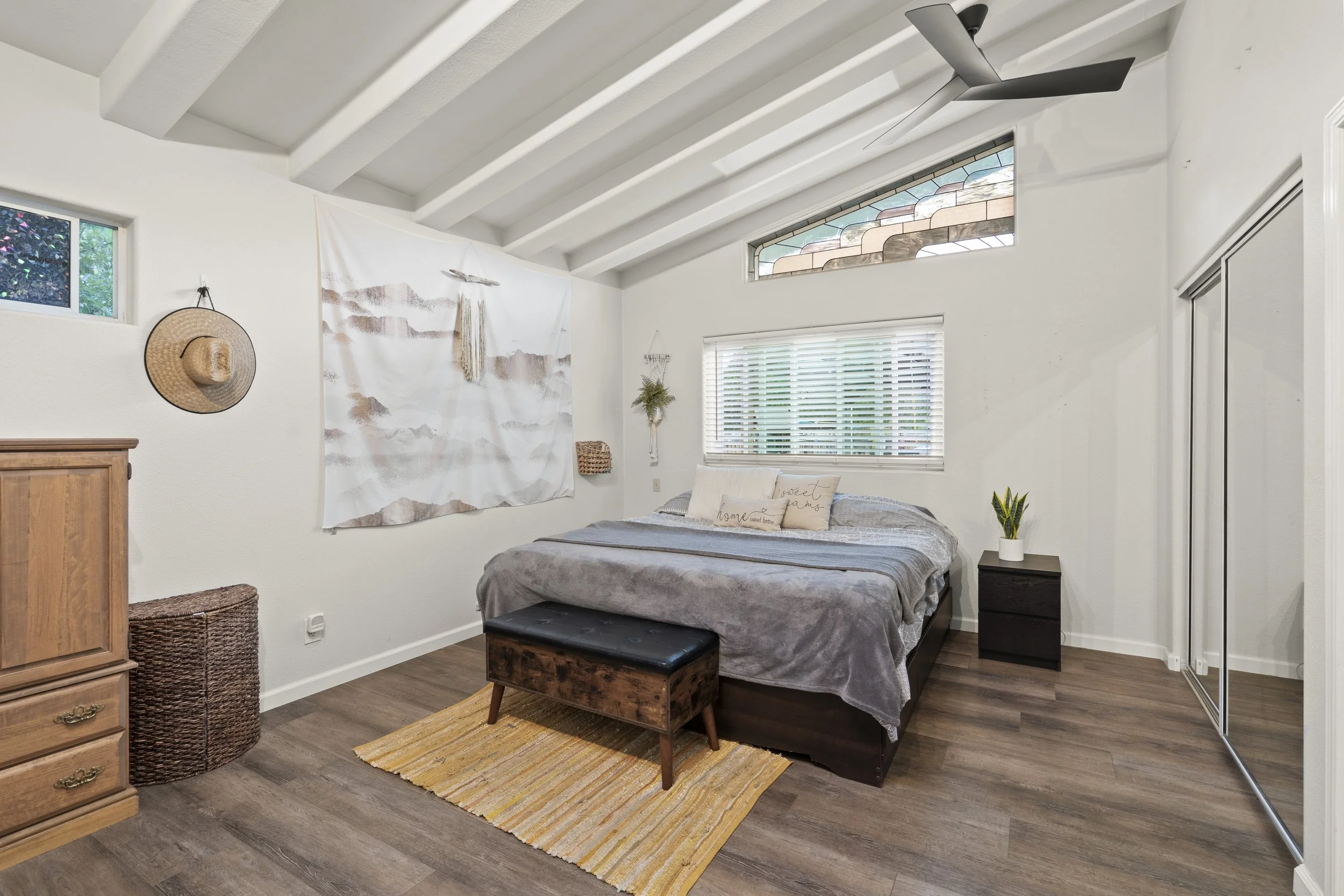 A bedroom with white walls and wood flooring, featuring a bed with gray blankets, a small wooden bench at the foot, and a nightstand with a plant. The room has a window with blinds, a ceiling fan, and decorative wall hangings.