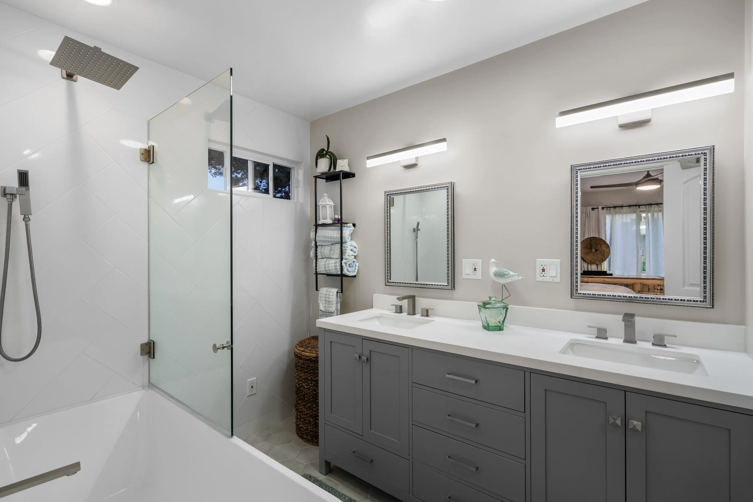 Bathroom with double vanity, gray cabinets, rectangular mirrors, modern light fixtures, and a walk-in shower with glass door, white tiles, and rainfall showerhead.