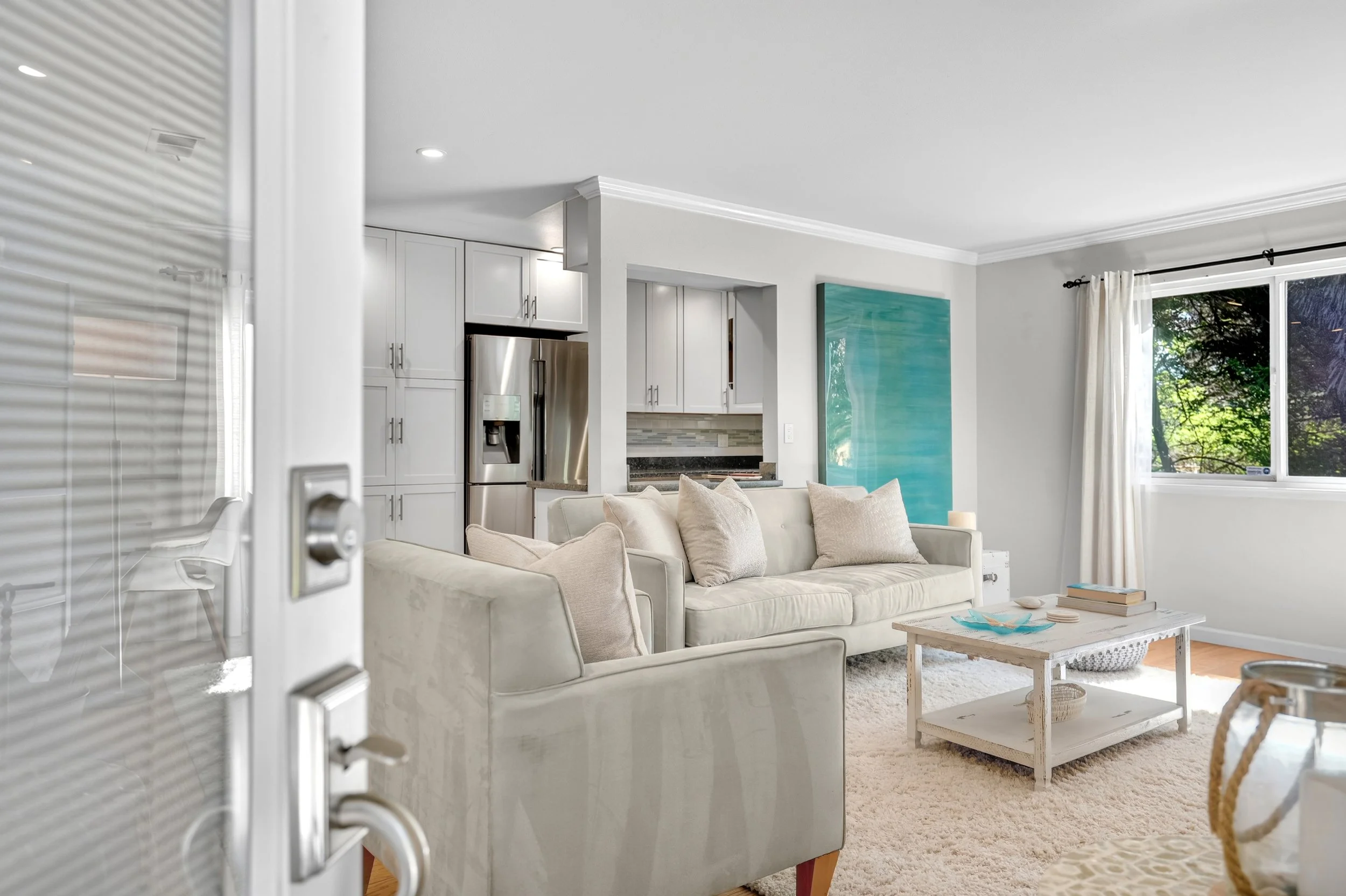 Living room with white couches, beige pillows, white coffee table, large window with white curtains, kitchen area with white cabinets and stainless steel refrigerator, large turquoise painting on the wall, and natural light.