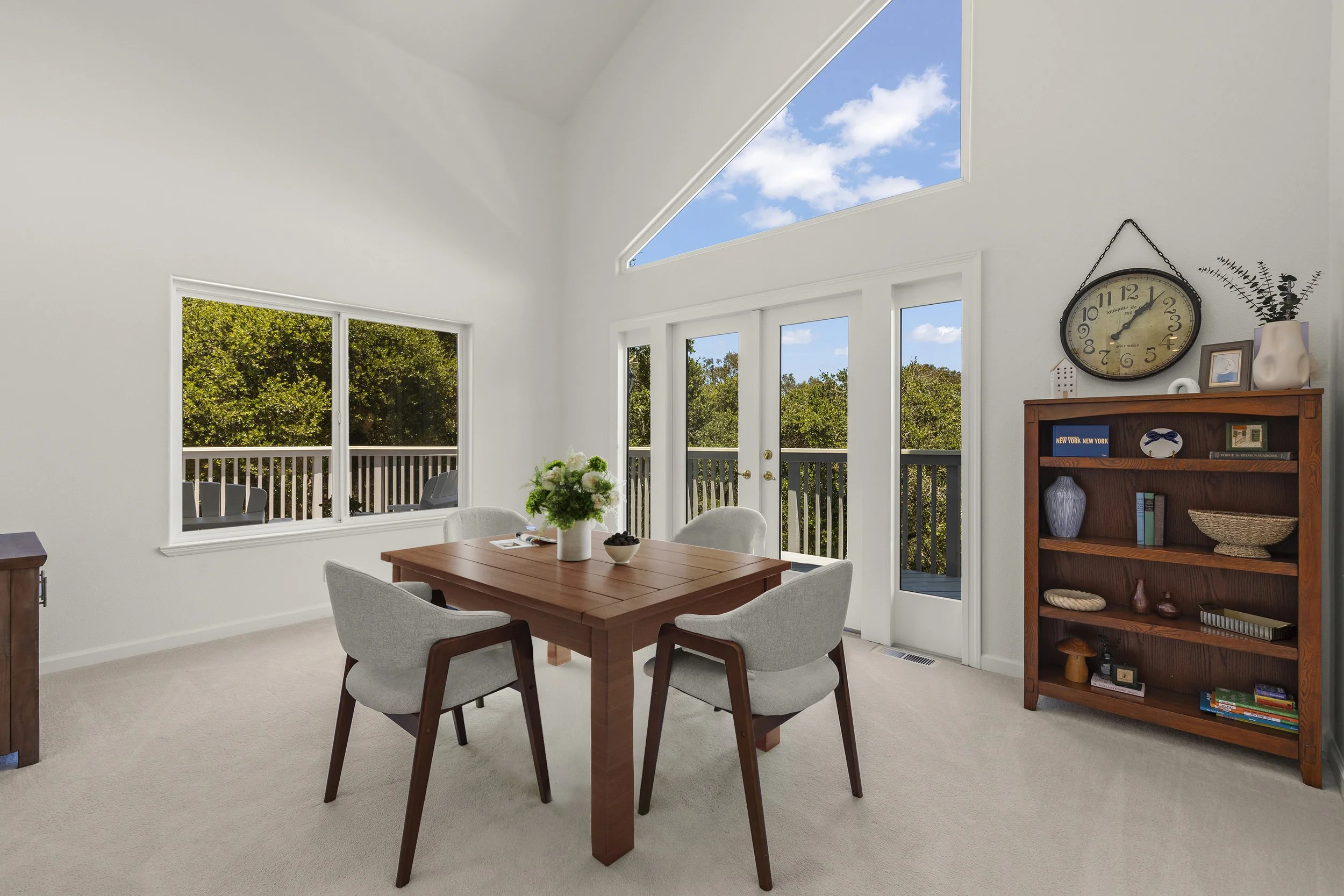 Bright dining room with a wooden table, four gray upholstered chairs, a vase of flowers, a wooden bookshelf, large windows, and a glass door leading to a balcony with trees outside under a blue sky with clouds.