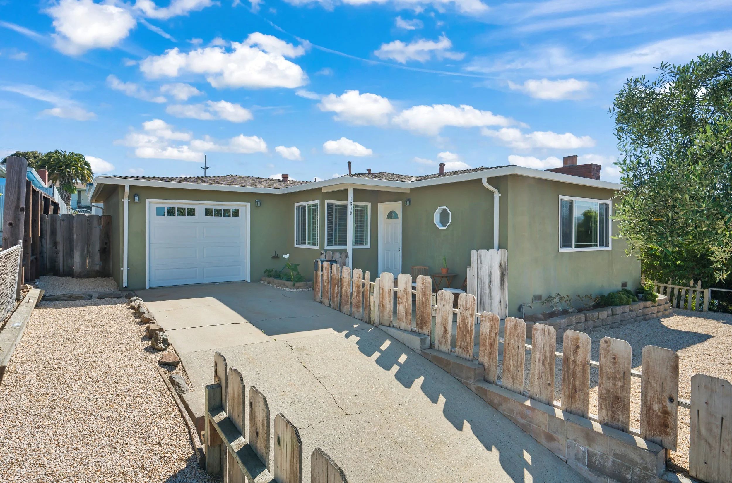 Single-story house with light green exterior, white garage door, and white front door. Small front yard with wooden fence, gravel, and concrete driveway. Bright blue sky with scattered clouds.