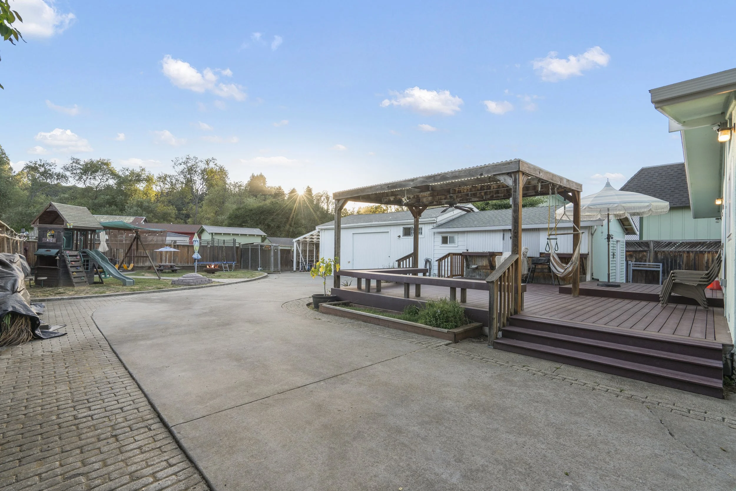 Backyard with a paved driveway, playset with slide, fire pit, raised wooden deck with swing chair and umbrella, surrounded by fence, trees, and houses, during sunset.