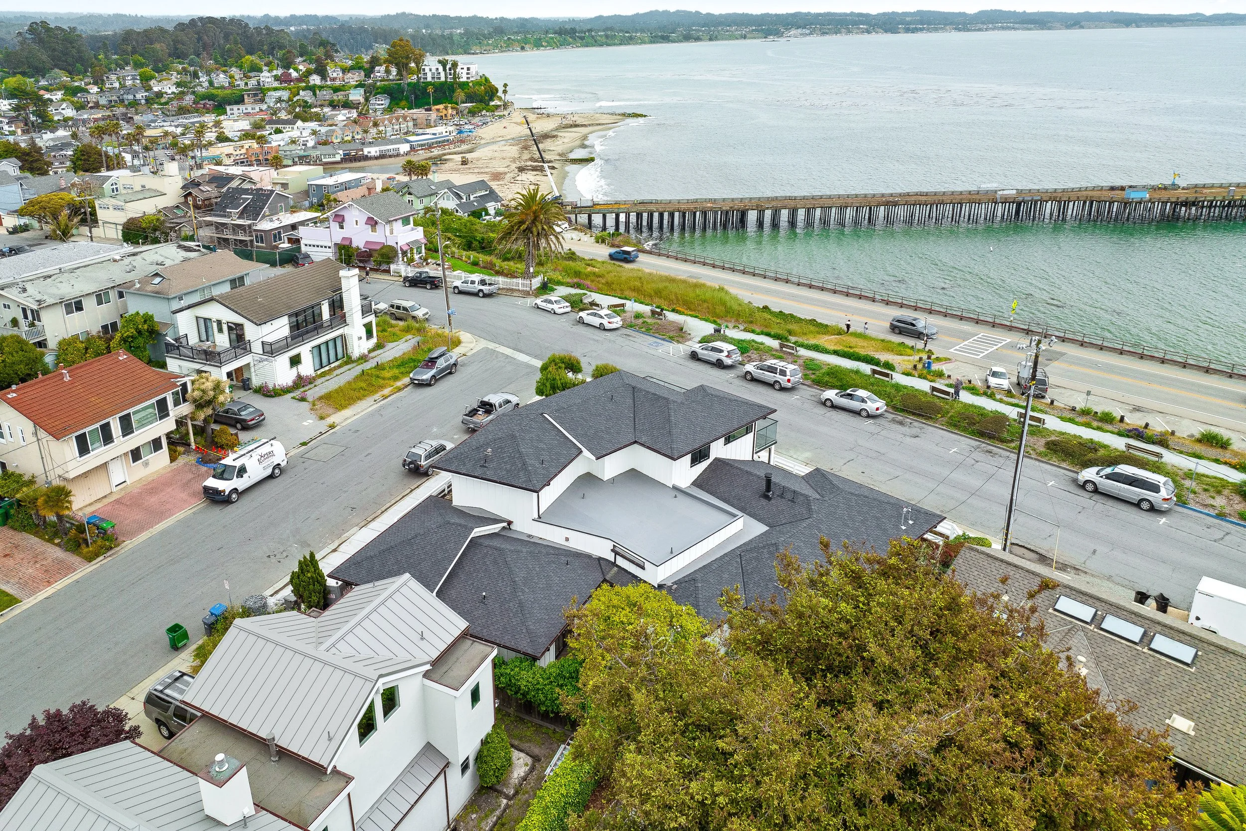 Aerial view of a coastal neighborhood with houses lining the street and a pier extending into the ocean, with a sandy beach and calm water. in Capitola.