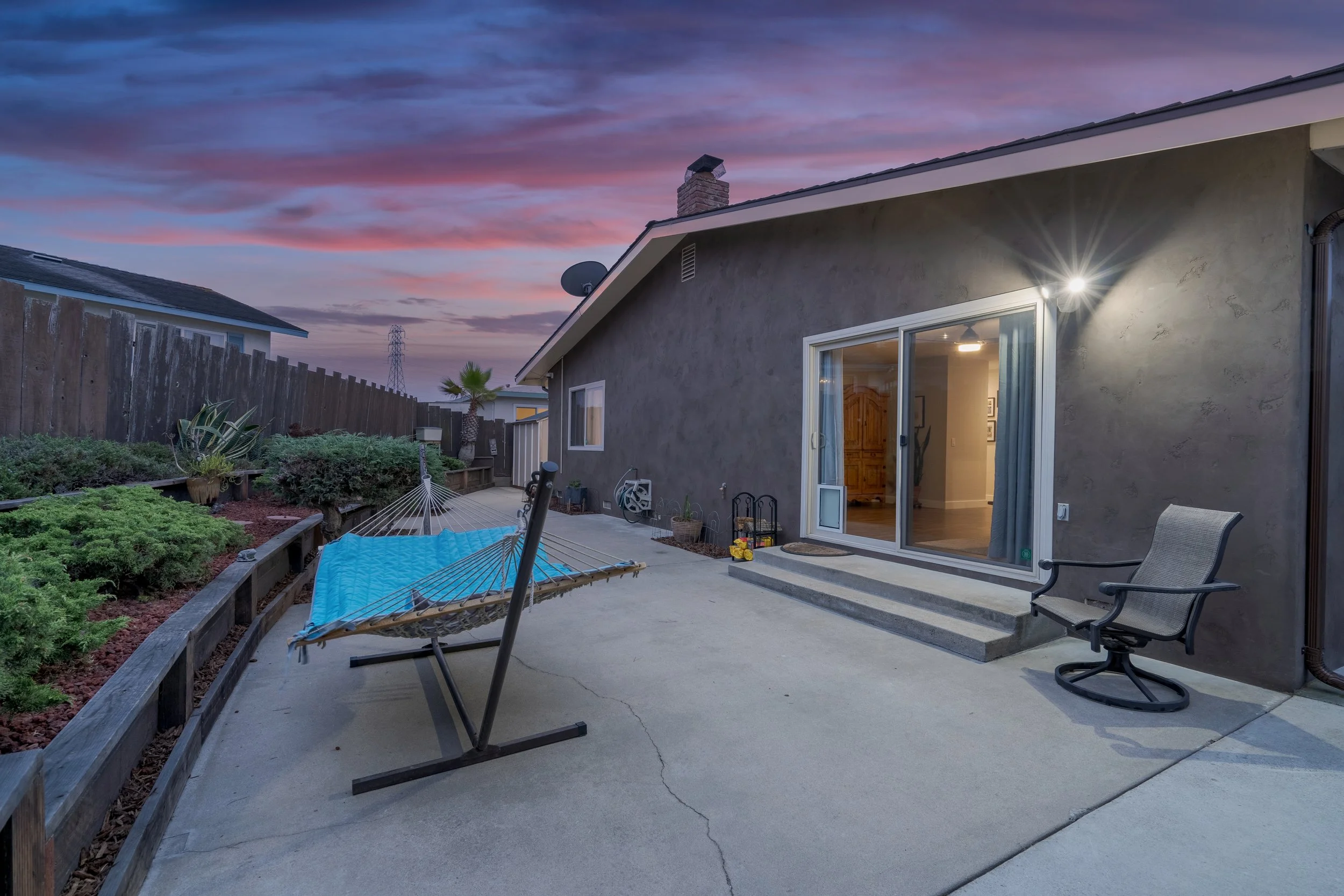 Backyard patio with a hammock, garden bed, outdoor chair, and a sliding glass door at dusk with a colorful sky.