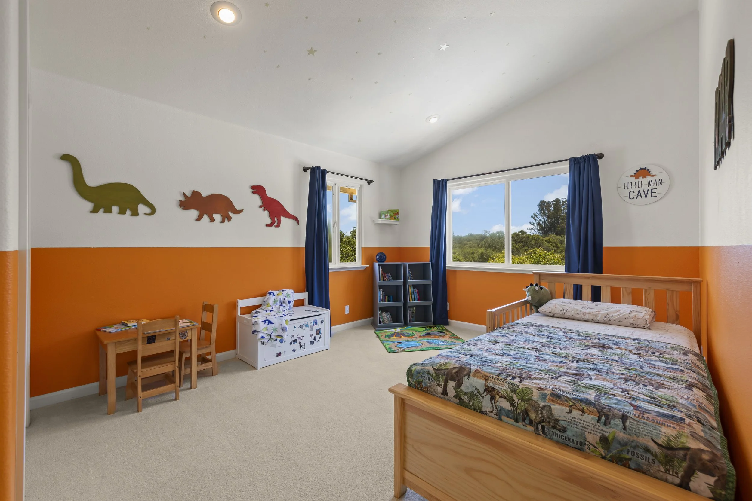 Children's bedroom with dinosaur-themed decor, a bed with dinosaur-themed bedding, a small table with chairs, a bookshelf, and windows with blue curtains showing trees outside.