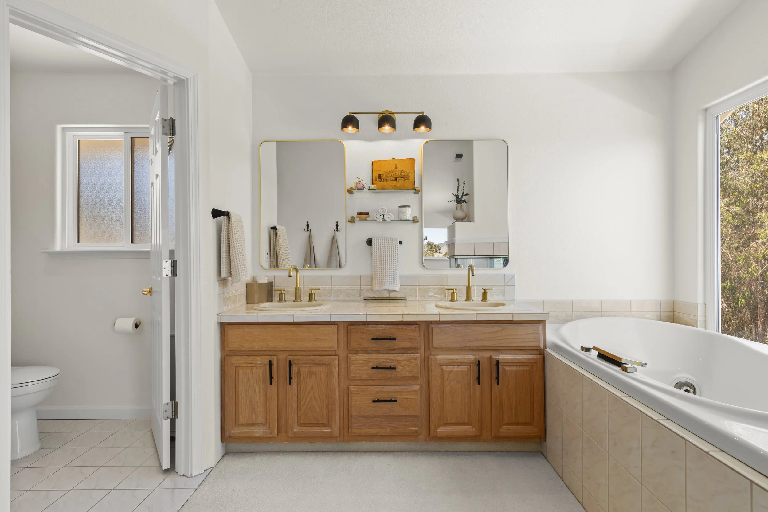 Bathroom with double vanity, two mirrors, gold faucets, wooden cabinets, a bathtub near a window, and a separate toilet room.