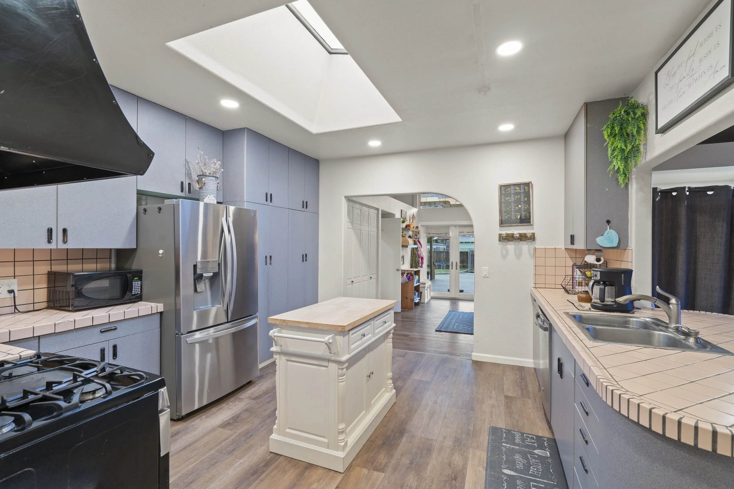 Kitchen with blue cabinets, pink tiled counters, stainless steel refrigerator, black microwave, stove, small white central island, and sliding glass doors.