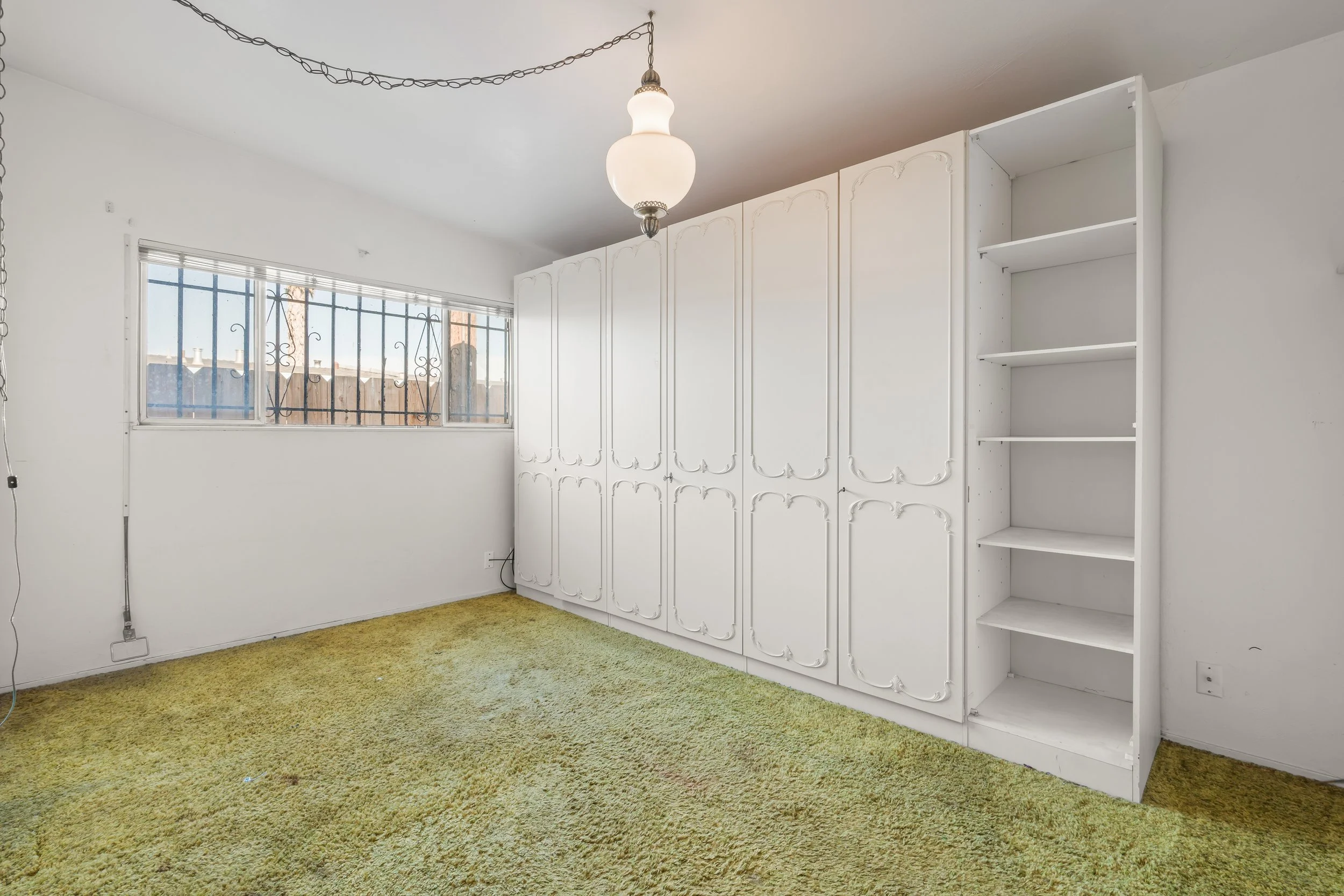 Empty room with white walls, green shag carpet, large window with metal bars, a ceiling light fixture, and built-in white cabinets and shelves in Salinas, Monterey county, CA.
