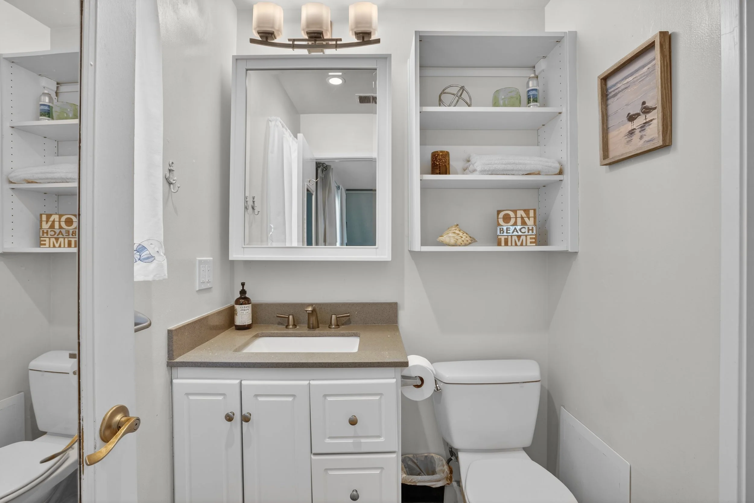 A small bathroom with a white vanity, beige countertop, and a setup with shelves above the toilet holding towels and decorative items. There's a mirror above the sink, a wall-mounted light fixture, and a framed seaside artwork. A partial view of a to