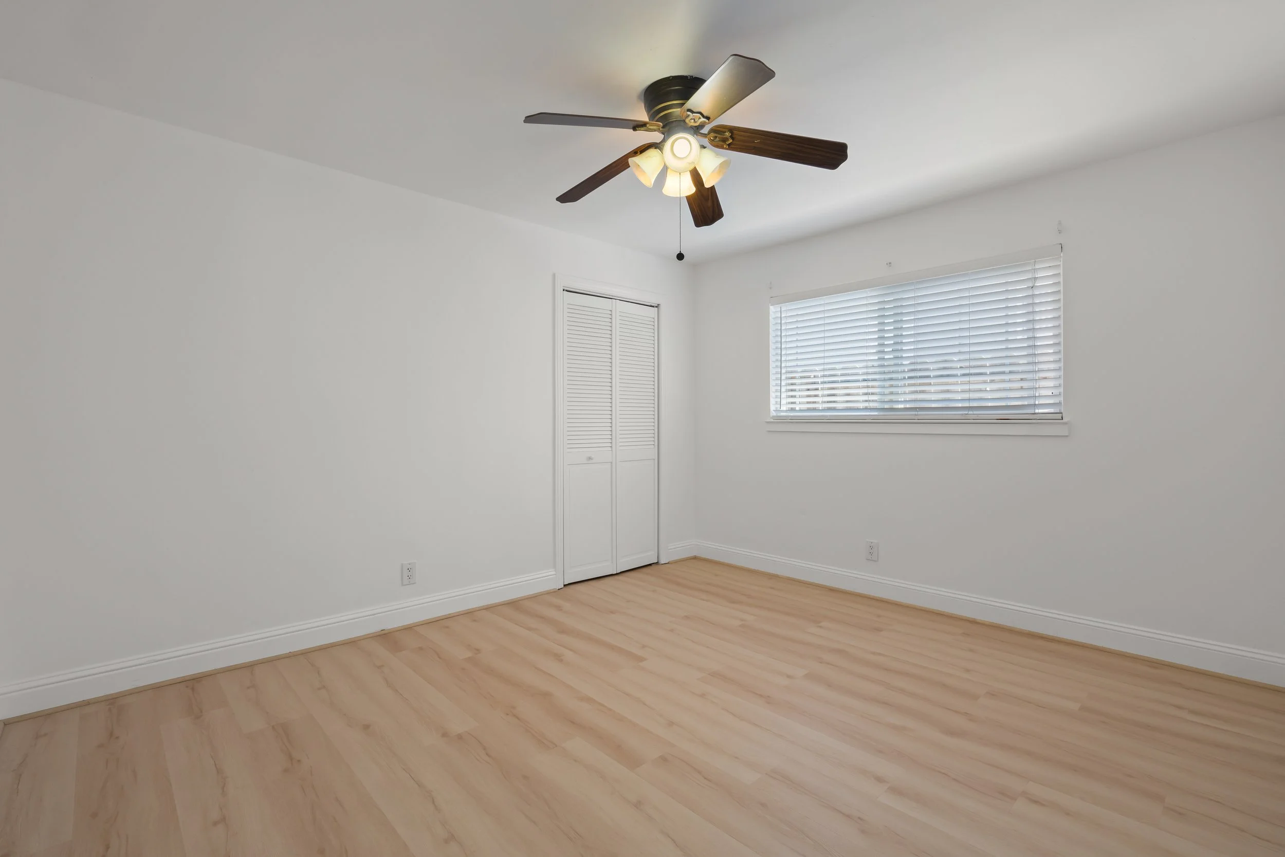 Empty room with light wood flooring, white walls, a window with blinds, a closet with louvered doors, and a ceiling fan with lights.