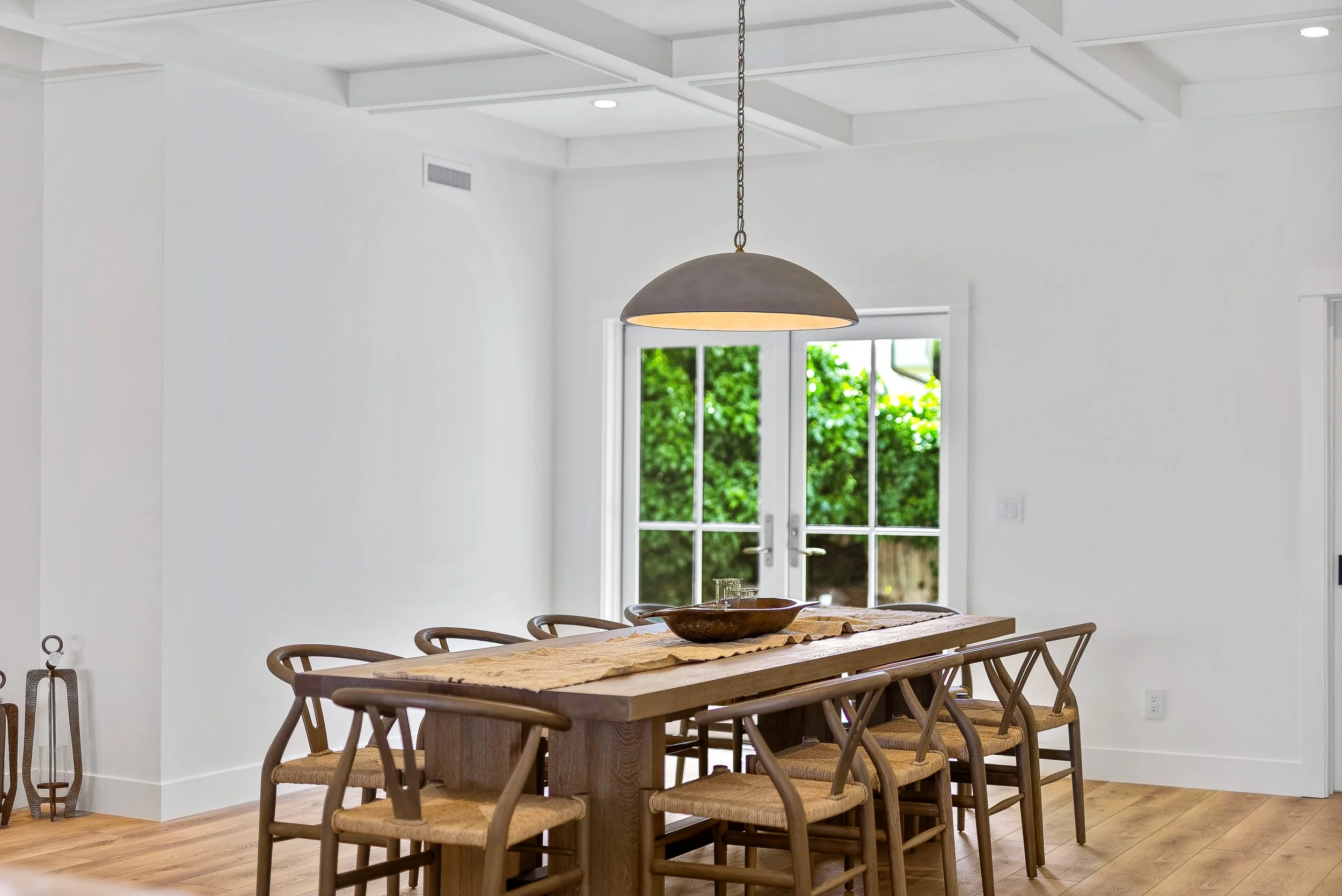 Dining room with a wooden table, eight chairs, a hanging light fixture, white walls, and glass doors leading to an outdoor garden in Capitola, California.