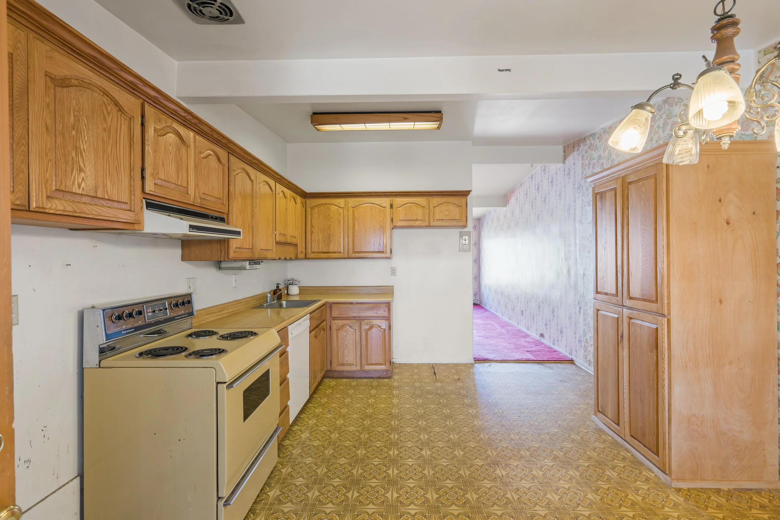 Kitchen with wooden cabinets, a yellow stove, and vintage patterned linoleum floor, leading to a dining area with floral wall decor.