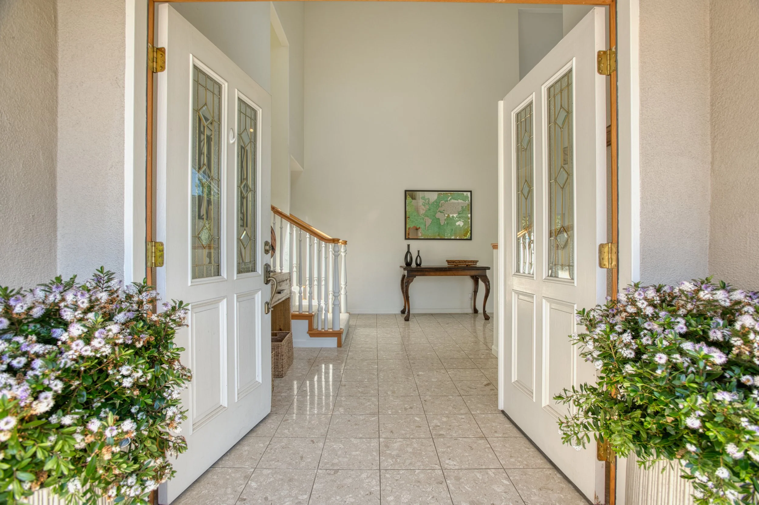 Open front door view of the entryway with flower pots on either side, tiled floor, staircase, and a table with decor in the background.