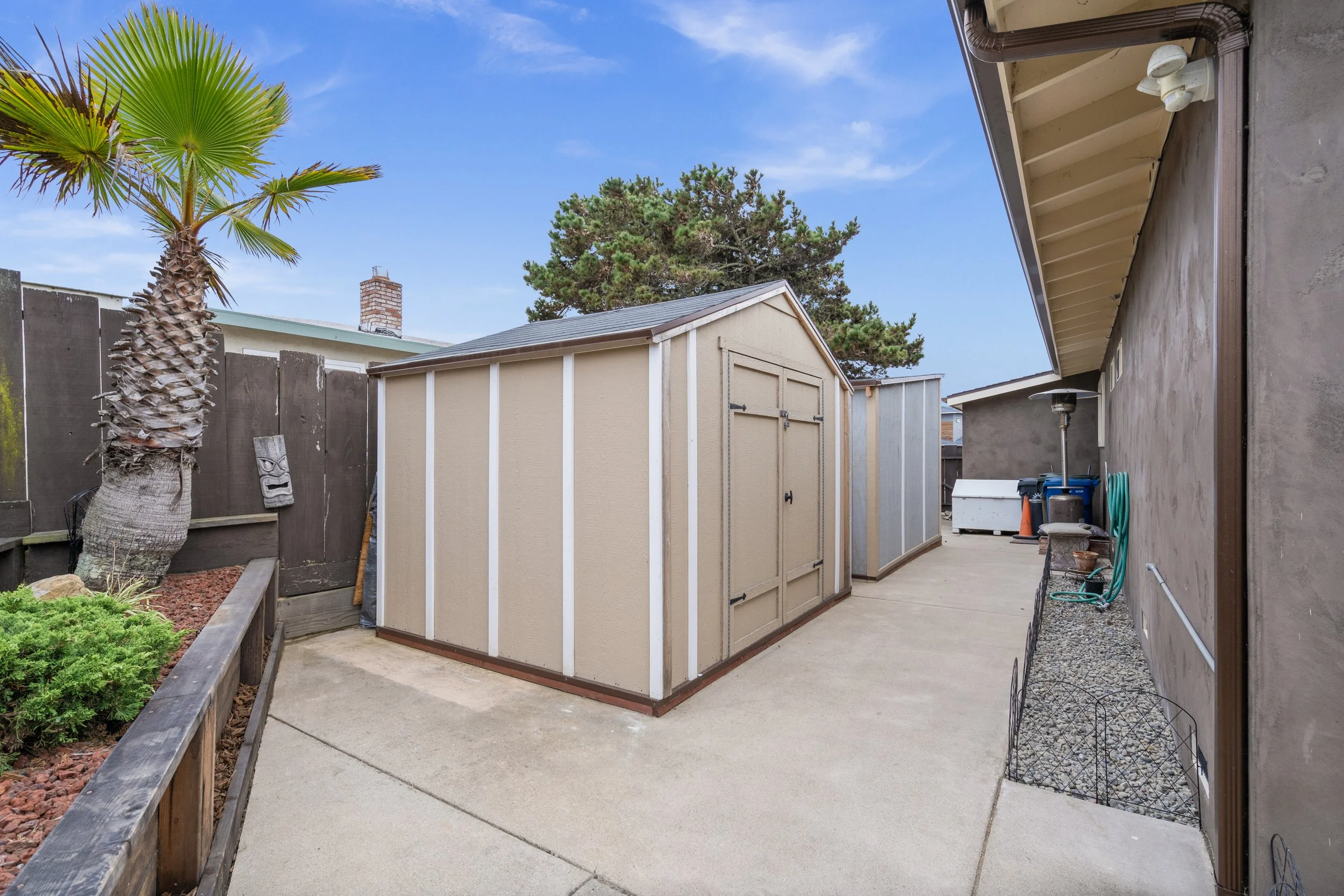 Backyard with two storage sheds, a palm tree, a wooden fence, and various outdoor items against the house wall.