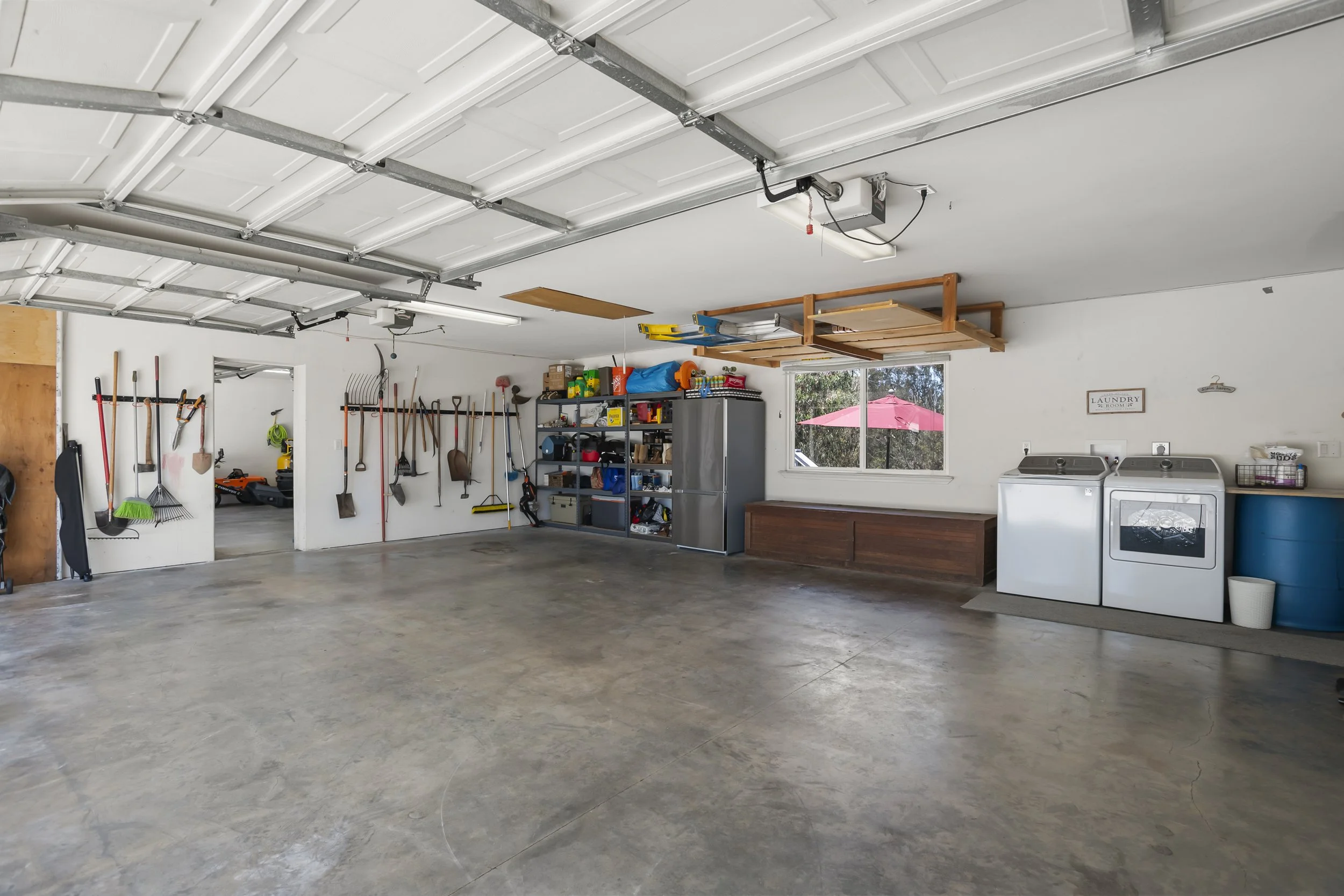 Empty garage with tools hanging on the wall, shelves with storage bins, a refrigerator, a washing machine and a dryer, and a window with a pink umbrella outside.