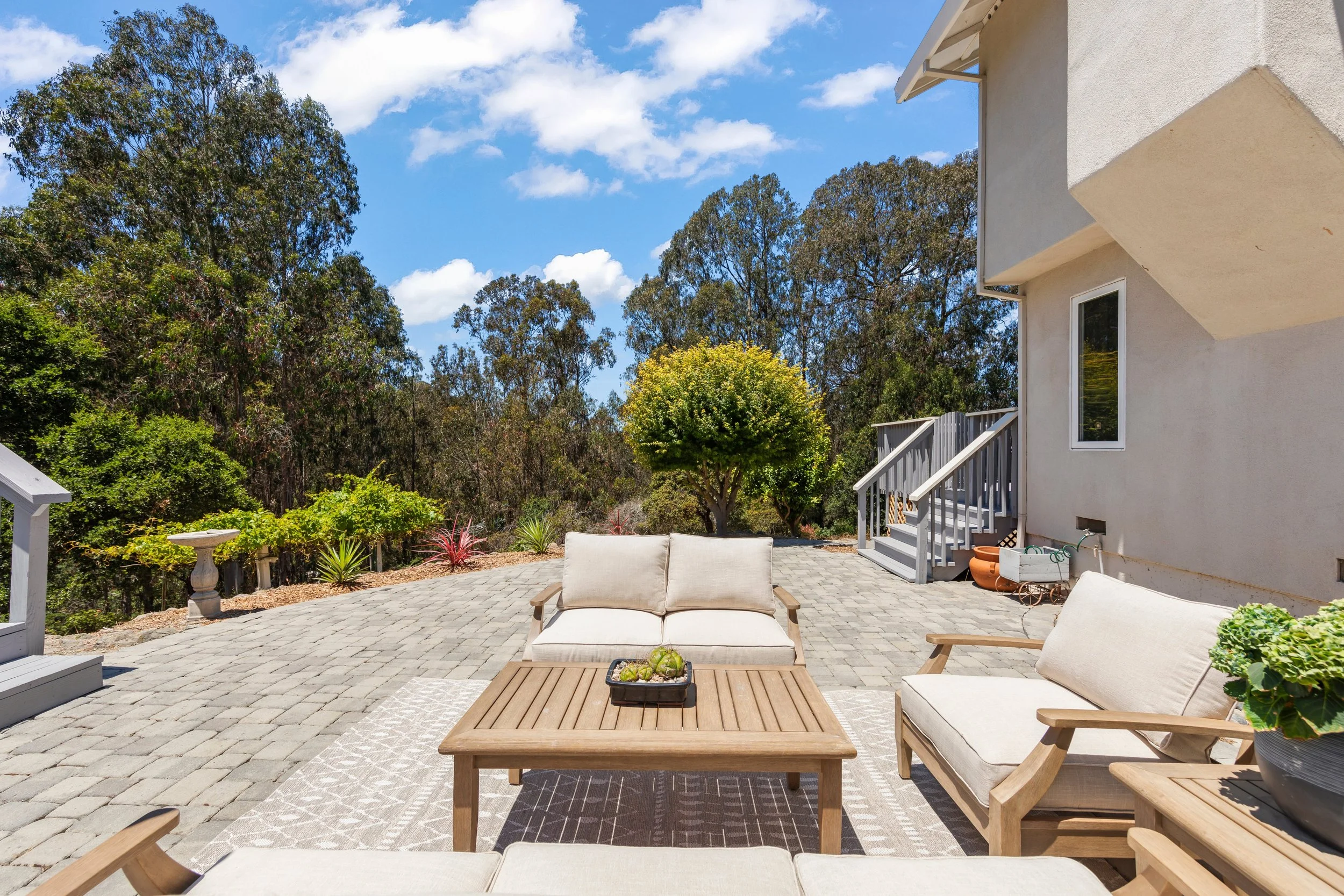 Outdoor patio area with cream-colored cushioned chairs and a wooden table on a stone paver surface, surrounded by trees and greenery under a partly cloudy sky.