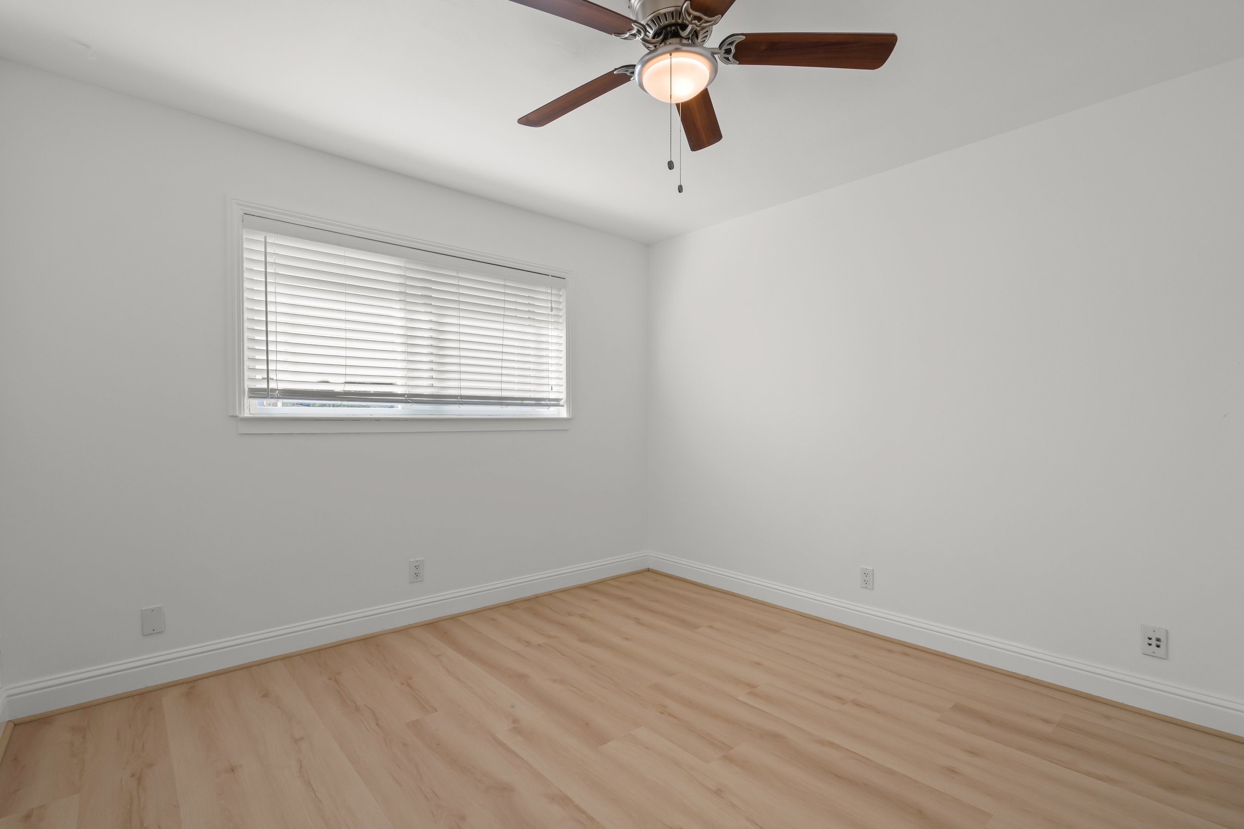 Empty room with white walls, light wood flooring, a window with white blinds, and a ceiling fan with wooden blades and a light fixture.