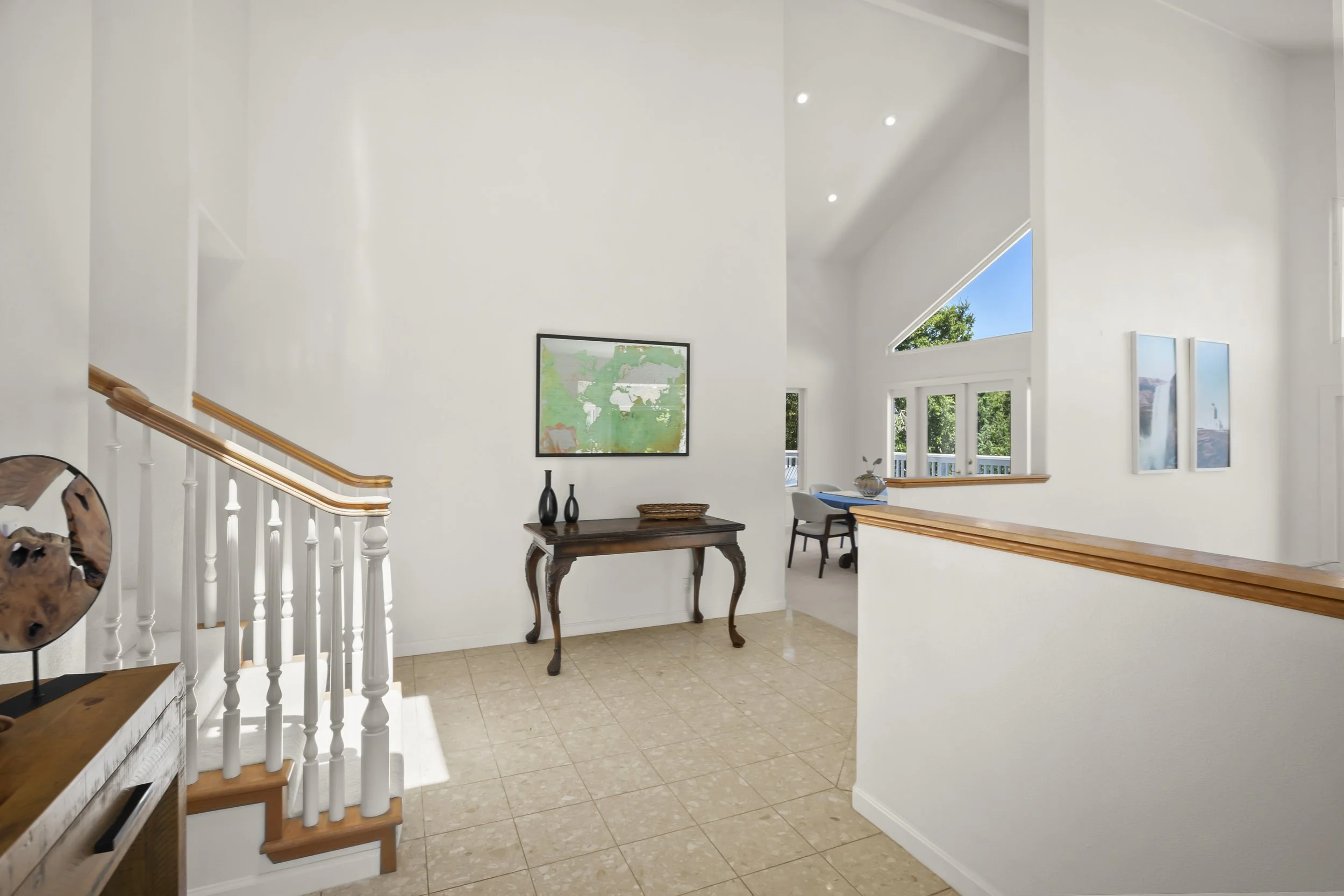 Spacious, well-lit interior entryway with white walls, tiled flooring, and a staircase with wooden handrail. Decor includes a dark wood console table with two black vases and a painting on the wall. In the background, there is a seating area with a desk near large windows and a sloped ceiling with recessed lighting.