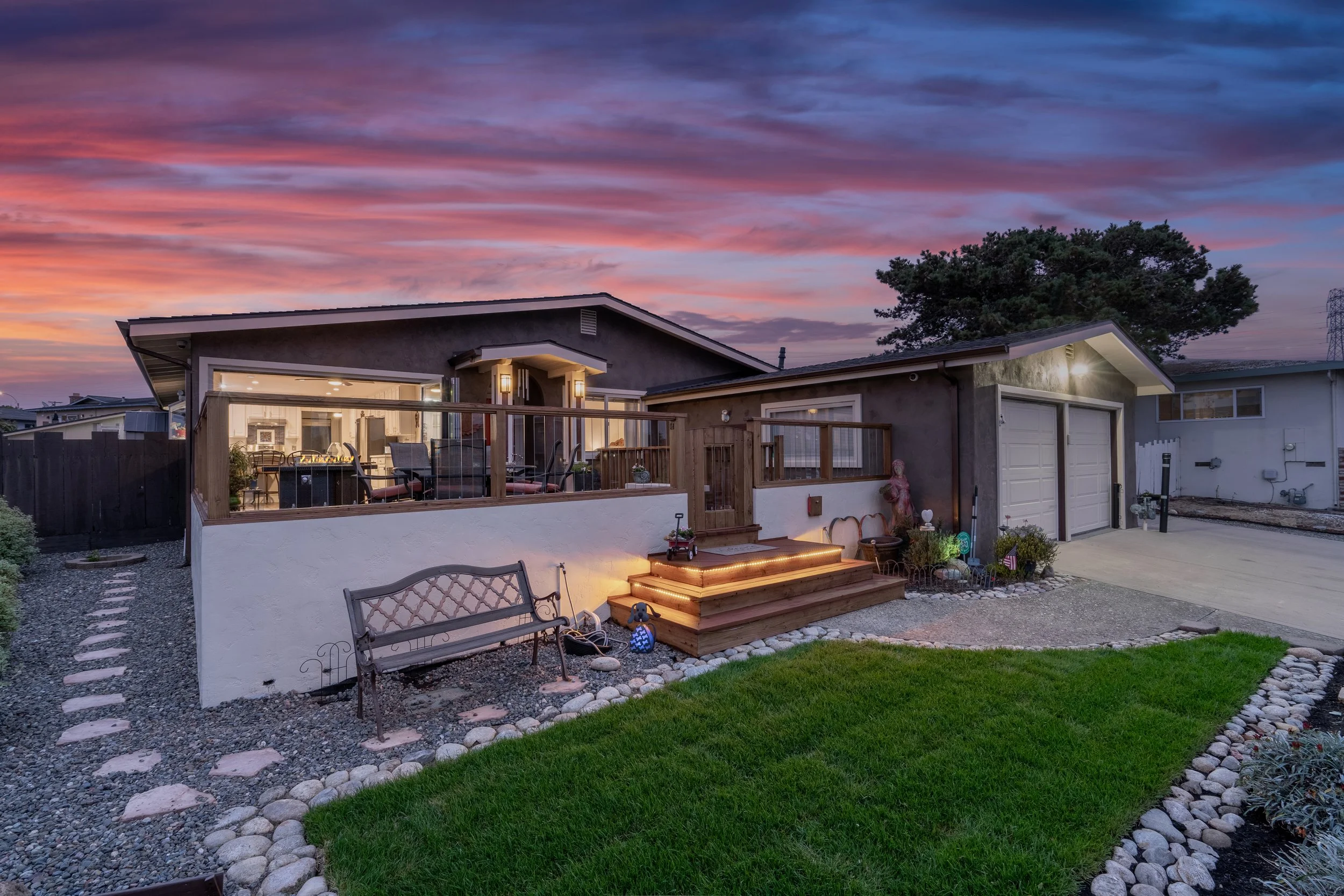 A house exterior during sunset with a wooden deck, outdoor seating, and a small garden with grass and stones.