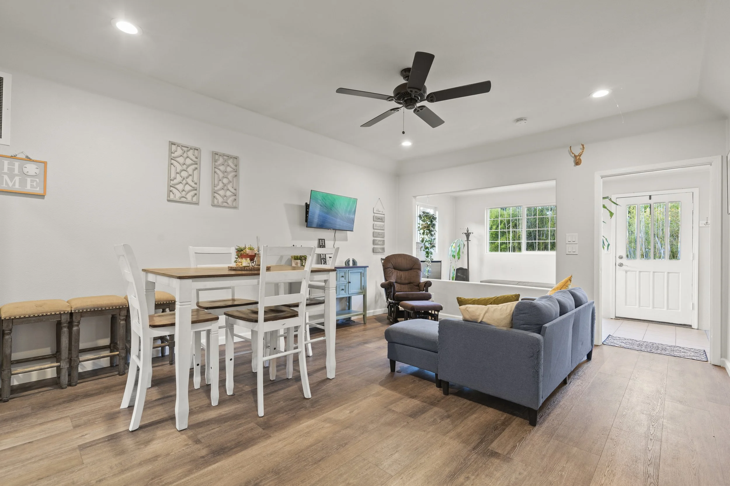 Living room with beige flooring, white walls, and a white ceiling. Includes a blue sofa with cushions, a brown recliner, a wall-mounted TV, a mix of white and wooden chairs around a dining table, and a small blue side table. Decor includes wall art, a deer head wall hanging, and several plants near the window.