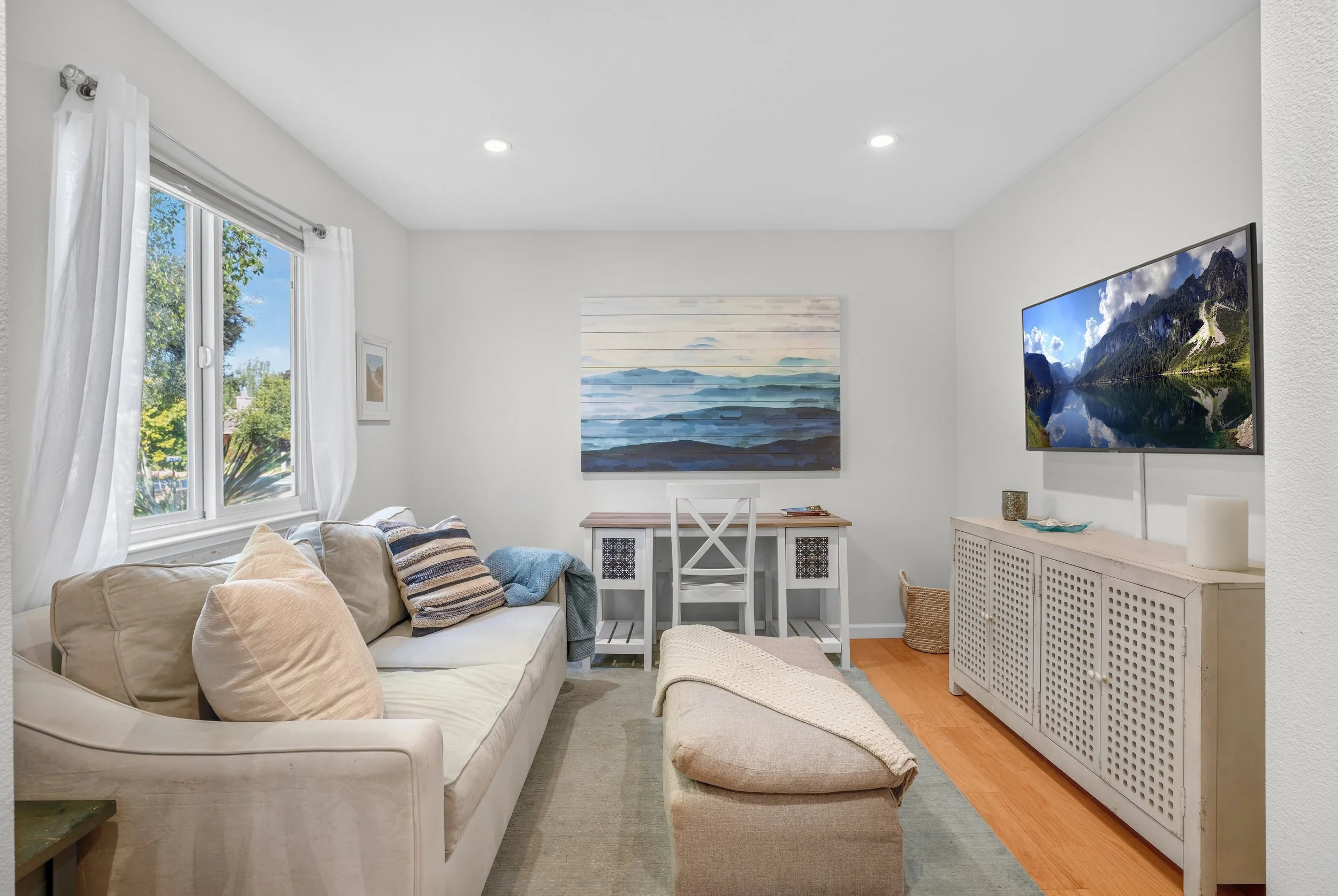 Living room with beige sofa, white walls, window with white curtains, wall art, and a mounted flat-screen TV showing a mountain lake scene in 1400 Dolphin Dr., Aptos CA.
