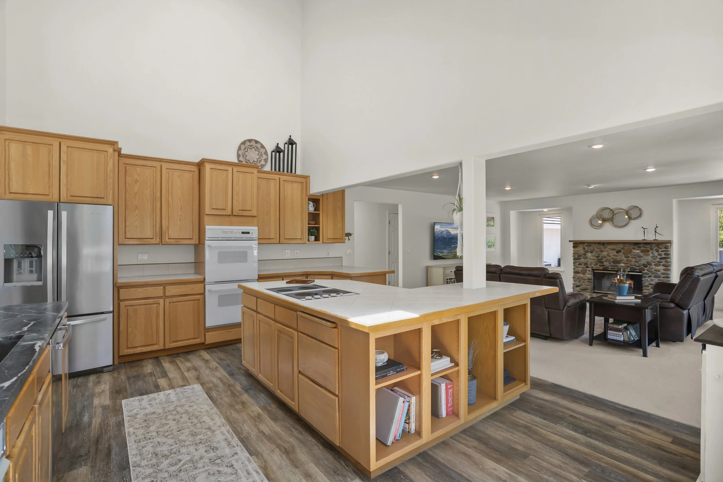 Open-concept kitchen with wooden cabinets, white appliances, and a central island with open shelving, leading into a living room with a stone fireplace and leather seating.