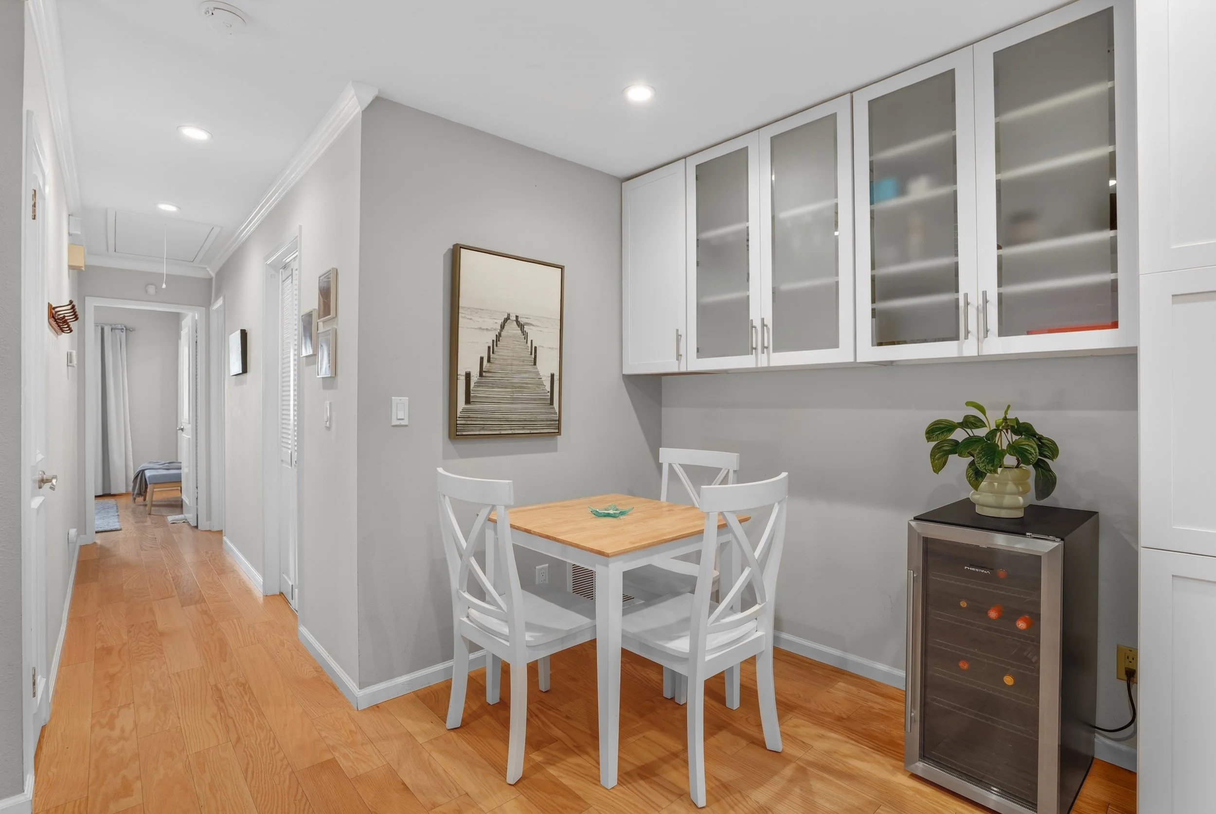 Dining area with a small wooden table, four white chairs, a potted plant, a wall-mounted cabinet with glass doors, and a picture of a pier over water.