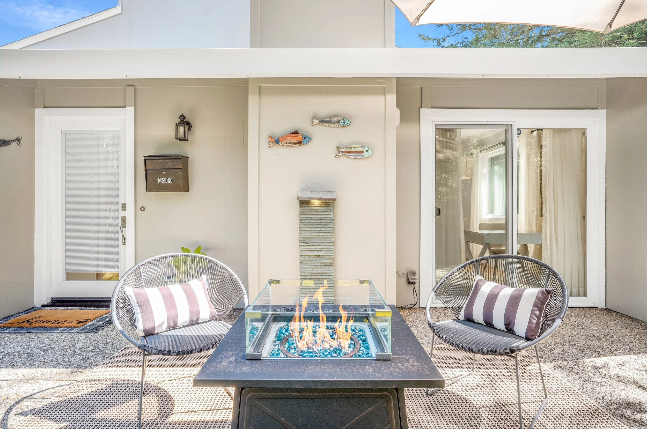 Outdoor patio with two black wire chairs, a fire pit table with glass enclosure, sliding glass door, and wall-mounted fish decorations.