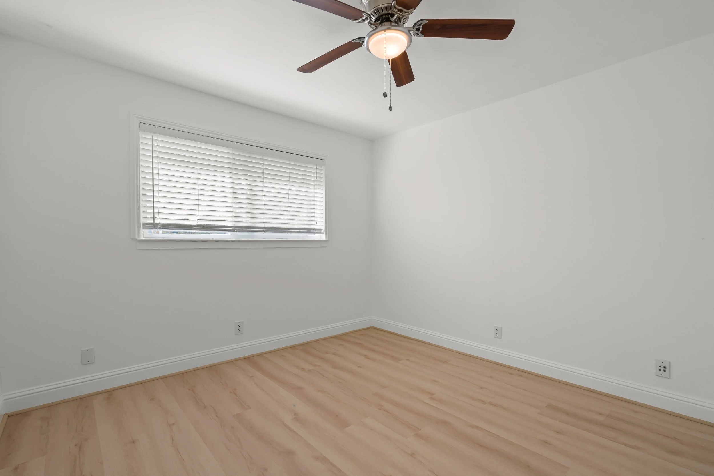 Empty room with white walls, wooden flooring, a window with white blinds, and a ceiling fan with a central light.