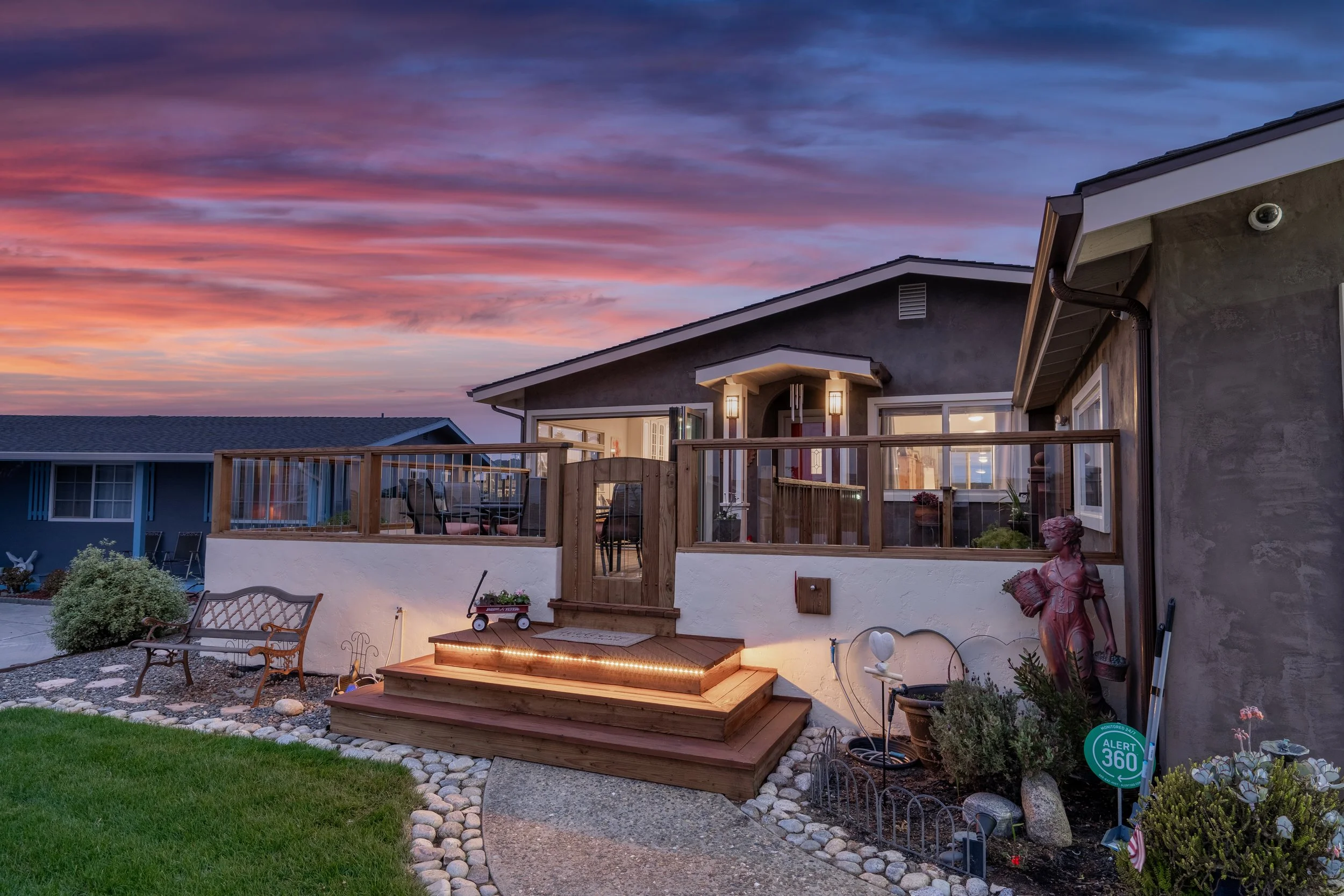 Backyard of a house during sunset with a wooden deck, outdoor seating, garden decorations, and a colorful sky.