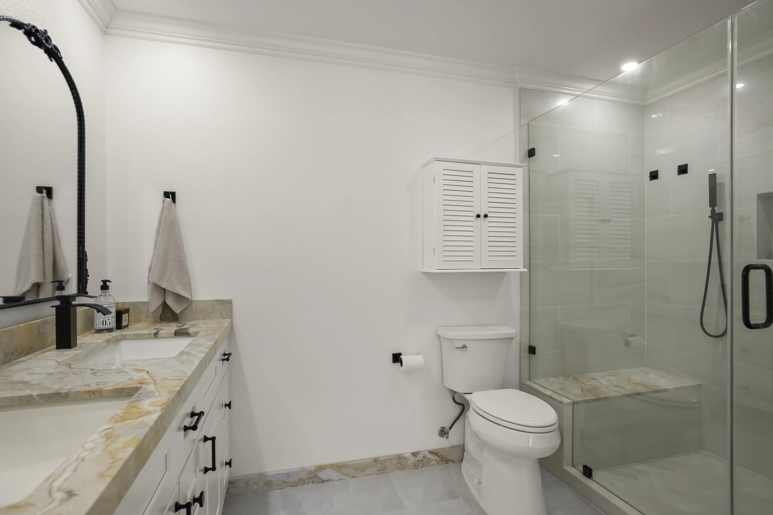Modern bathroom with white walls, marble countertop, double sinks, black fixtures, beige towels, a toilet, wall-mounted cabinet, and a glass-enclosed shower with a tiled bench.