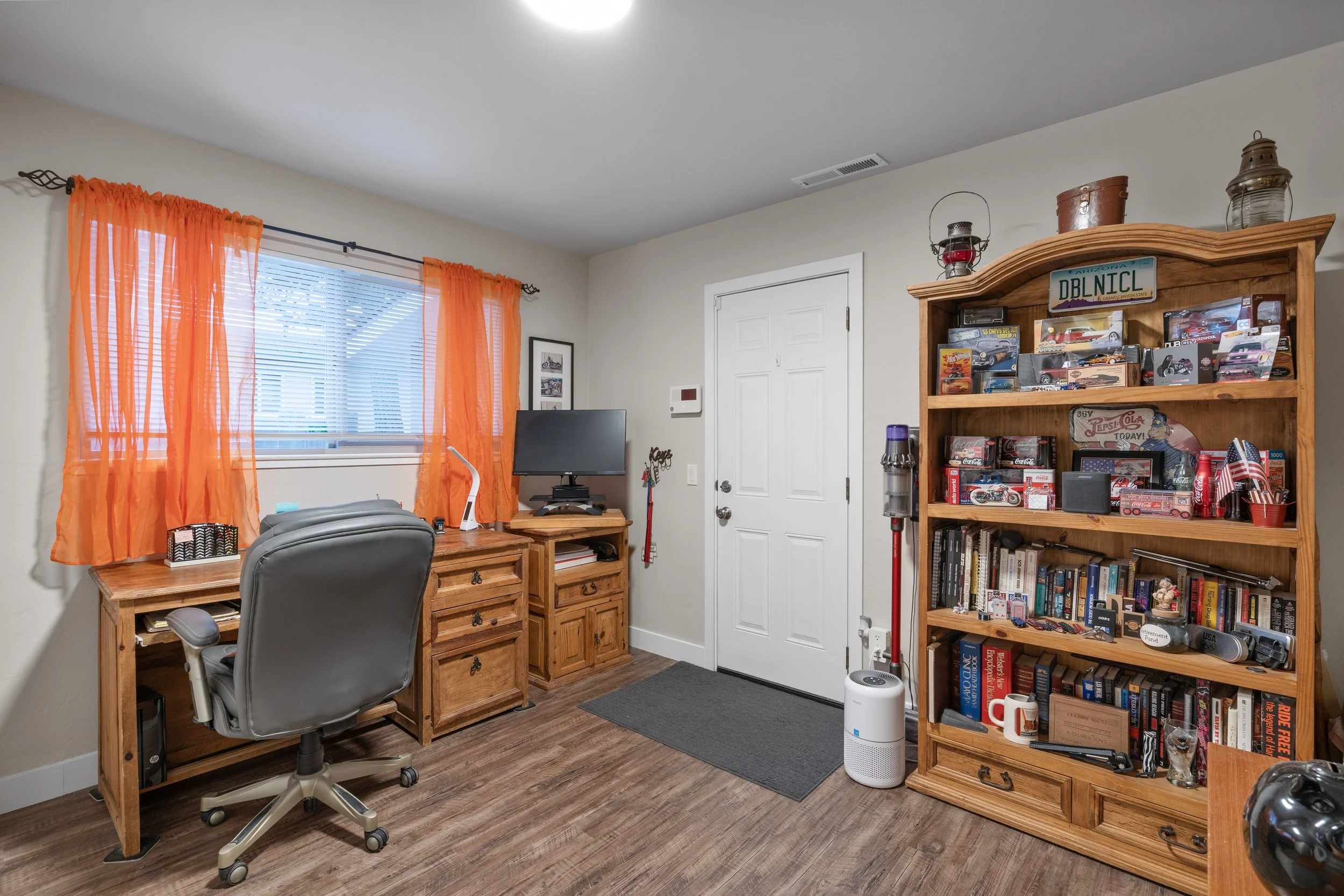 An office or study room with a wooden desk, a gray office chair, a computer monitor, and a wooden bookshelf filled with books and collectibles. The room has orange curtains on a window, a gray door, and a small gray mat in front of the door. There is