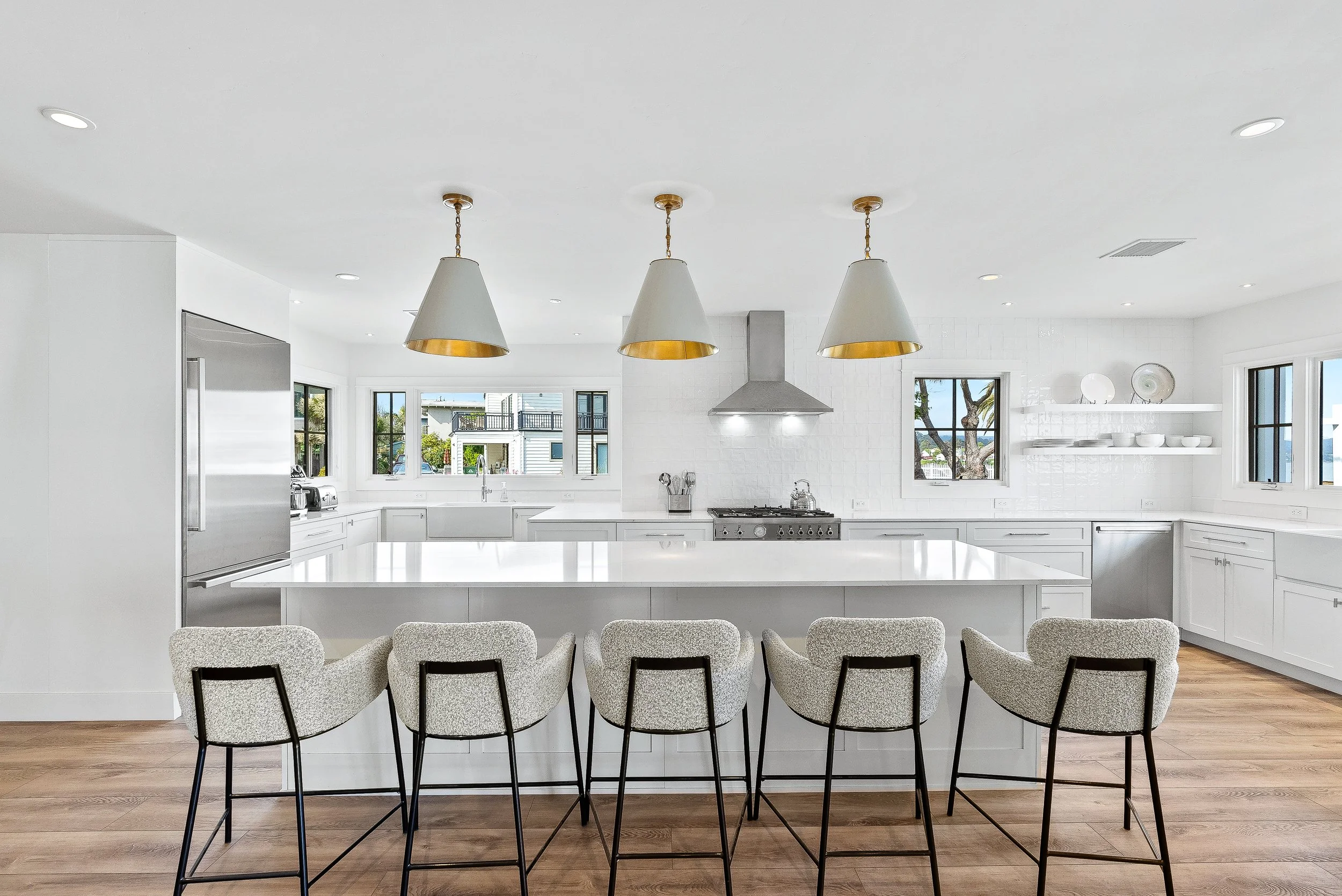 Bright, modern white kitchen with a large island, five beige barstools, stainless steel appliances, and pendant lights, with windows showing neighborhood houses outside.