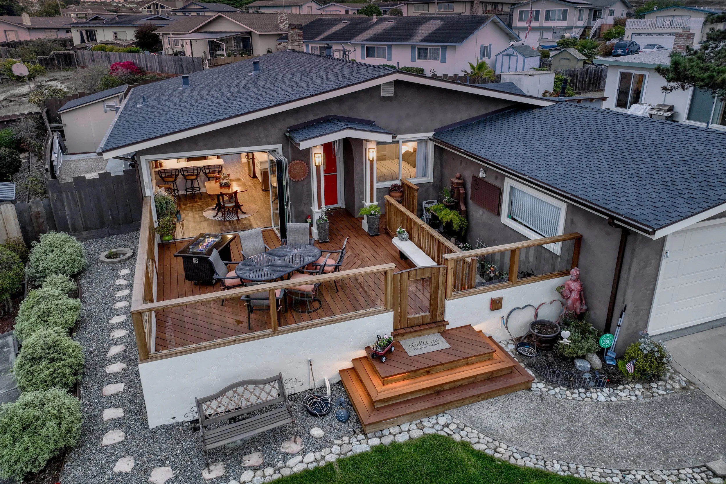 A backyard patio area with outdoor furniture and a wooden deck, adjacent to a house with large windows, in a suburban neighborhood.