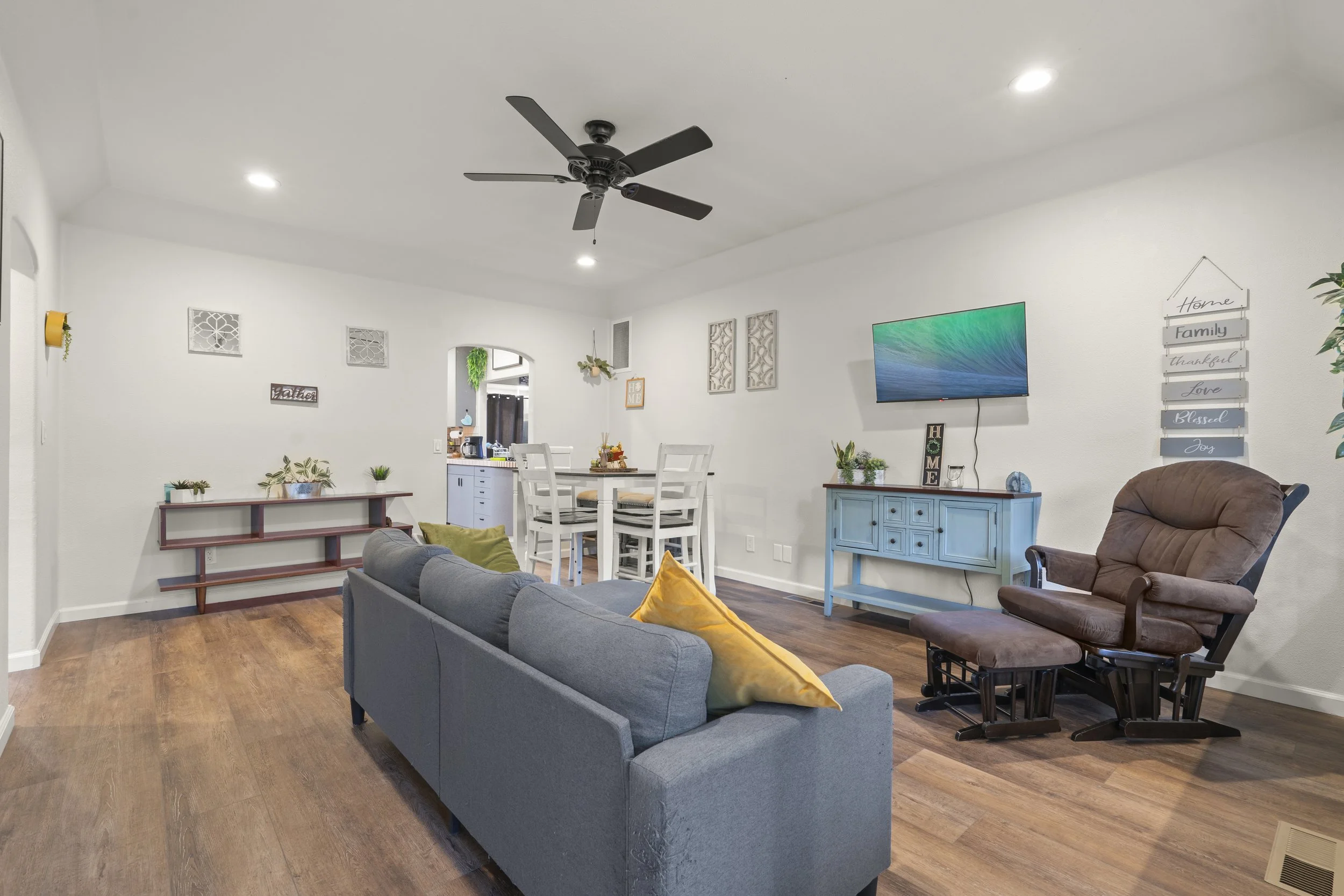 Living room with a gray sofa, a brown recliner, a blue console table, a wall-mounted TV, and a dining area in the background. Decor includes wall art, plants, and pillows, with wood flooring and ceiling lights.