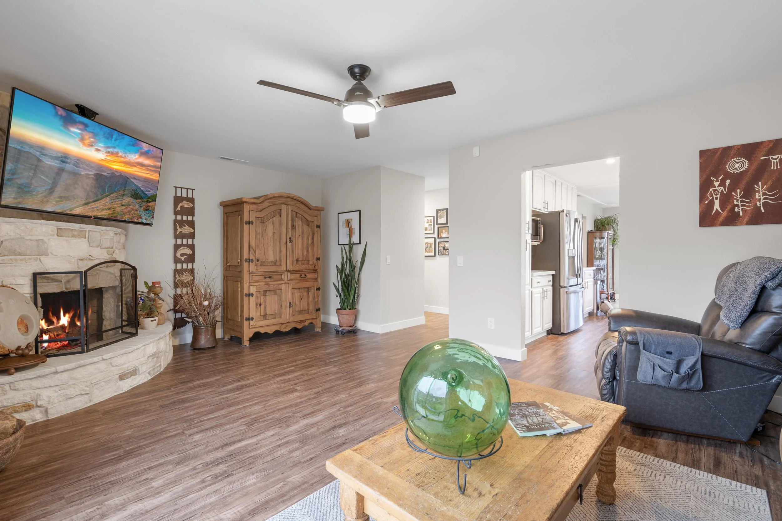 Living room with a stone fireplace, mounted TV, wooden furniture, ceiling fan, leather armchair, and a wooden coffee table with a green glass globe and magazines, with a view into a kitchen.