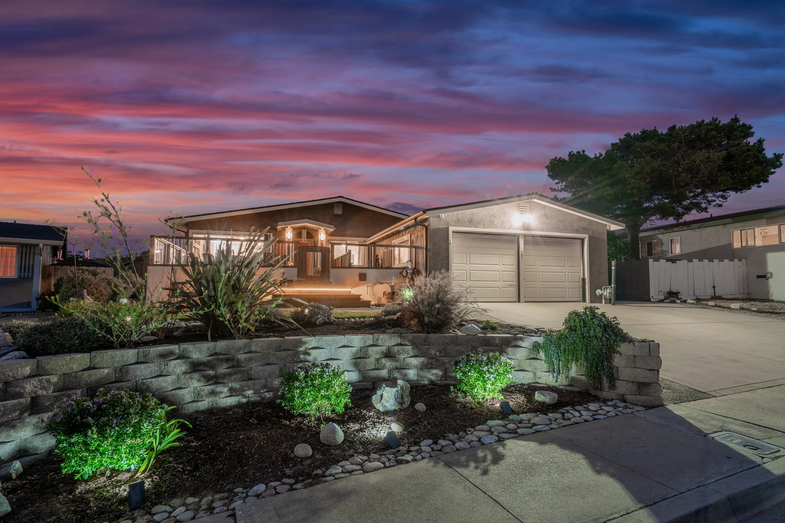 A single-story gray house with a two-car garage, illuminated exterior lights, a wooden deck, and a landscaped front yard with plants, rocks, and mulch, under a sunset sky with colorful clouds.