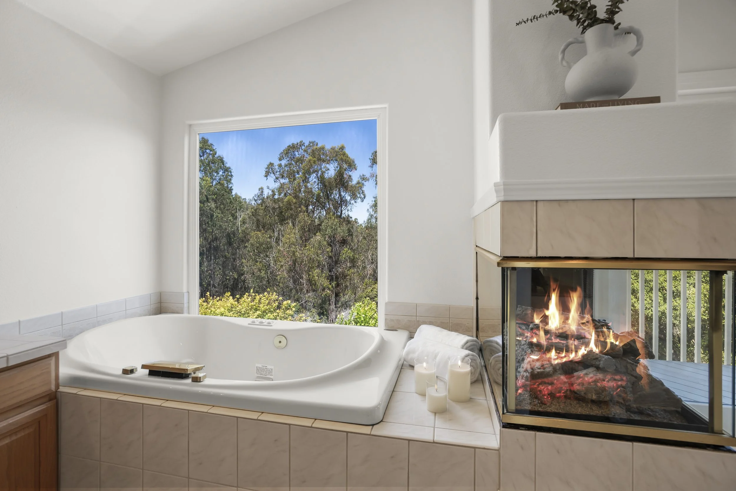 A bedroom with a large bathtub near a window with a view of trees, candles, folded towels, and a lit fireplace.