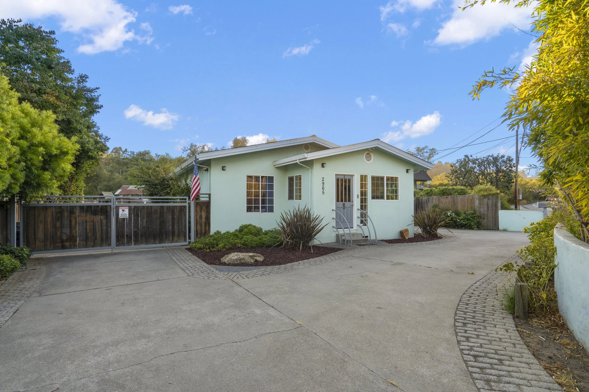 A light green house with white trim, a small front porch, and a American flag near the gate. The house is surrounded by greenery and a driveway with a curved pattern of concrete and bricks.