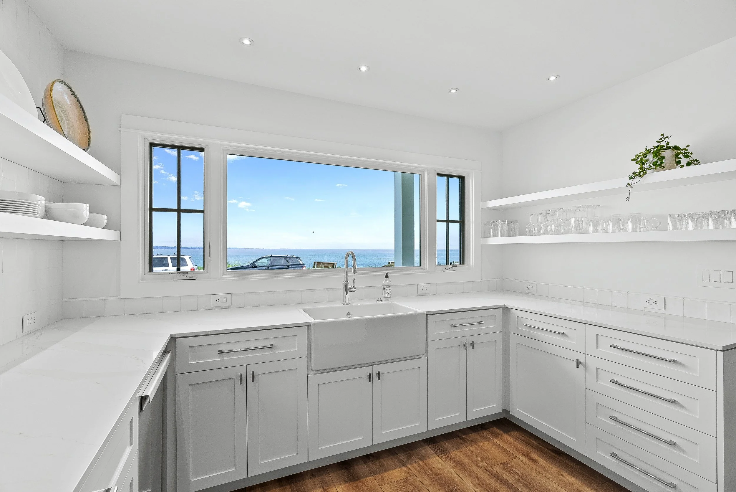 Bright white kitchen with large window overlooking a body of water and sky, white cabinets, open shelves with glassware and plates, wooden floor in Capitola, California.
